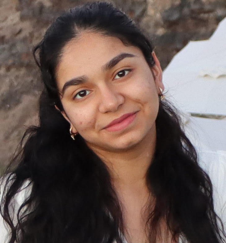 Close-up of a young woman with long dark hair, smiling softly, outdoors near water and rocks.