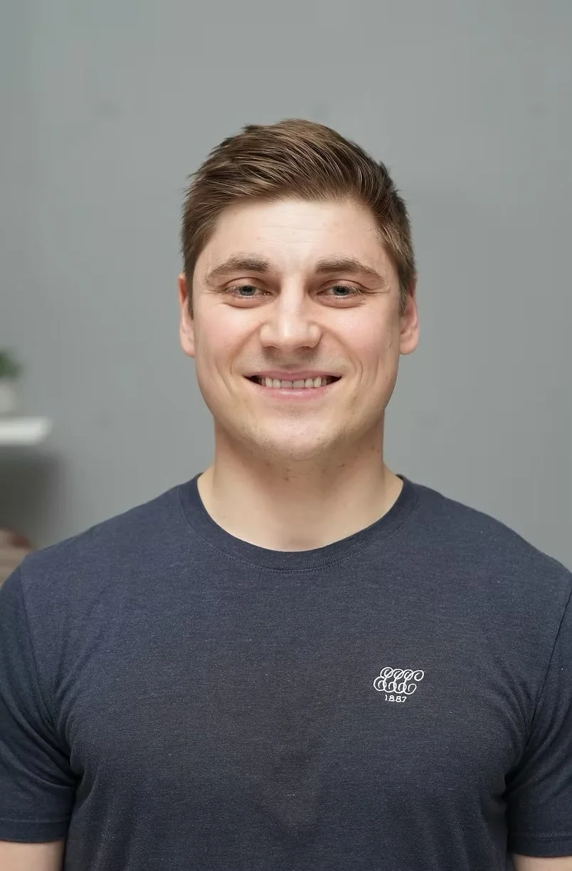 A young man with light skin, brown hair styled back, smiling, wearing a dark t-shirt with a small logo and the year 1887 on the chest, standing indoors against a light gray wall with a blurred background.