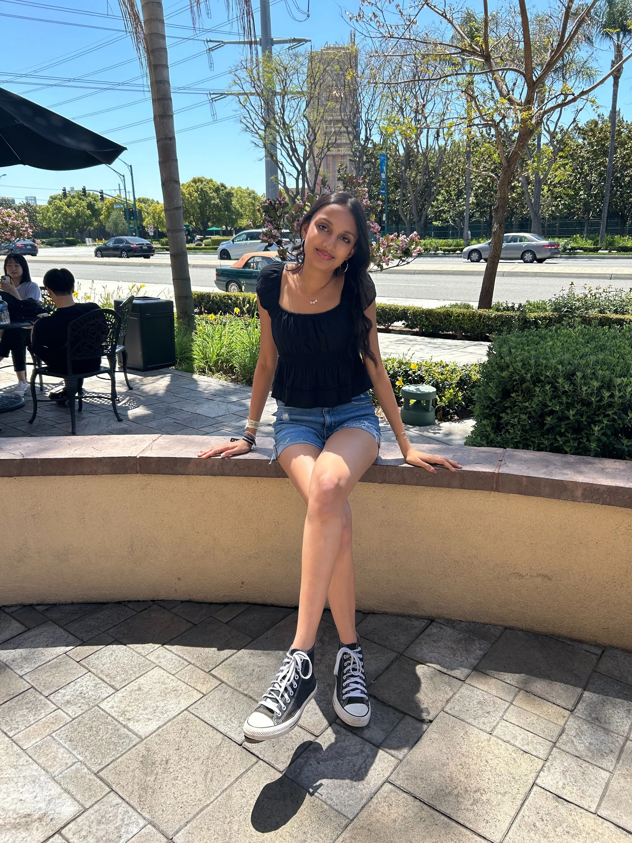 Young woman sitting on a curved outdoor wall, wearing a black top, denim shorts, and black sneakers with white laces, smiling at the camera with cars and trees in the background.