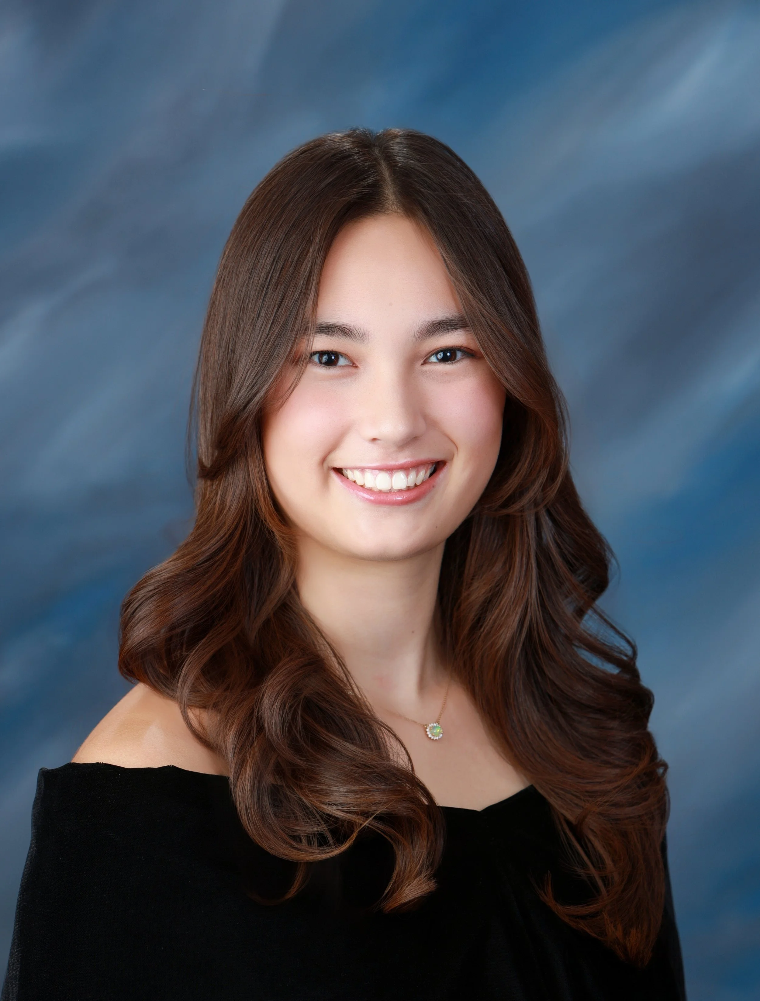 A young woman with long, wavy brown hair and a bright smile, wearing a black off-the-shoulder top and a necklace with a green pendant, against a blue and gray blurred background.