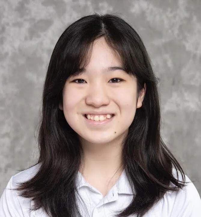 A young woman with shoulder-length dark hair, smiling, wearing a white collared shirt, standing against a textured gray background.