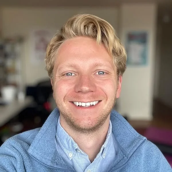 A young man with blonde hair, blue eyes, and a wide smile, wearing a light blue collared shirt and a blue zip-up sweater. He is in an indoor setting with a blurred background.