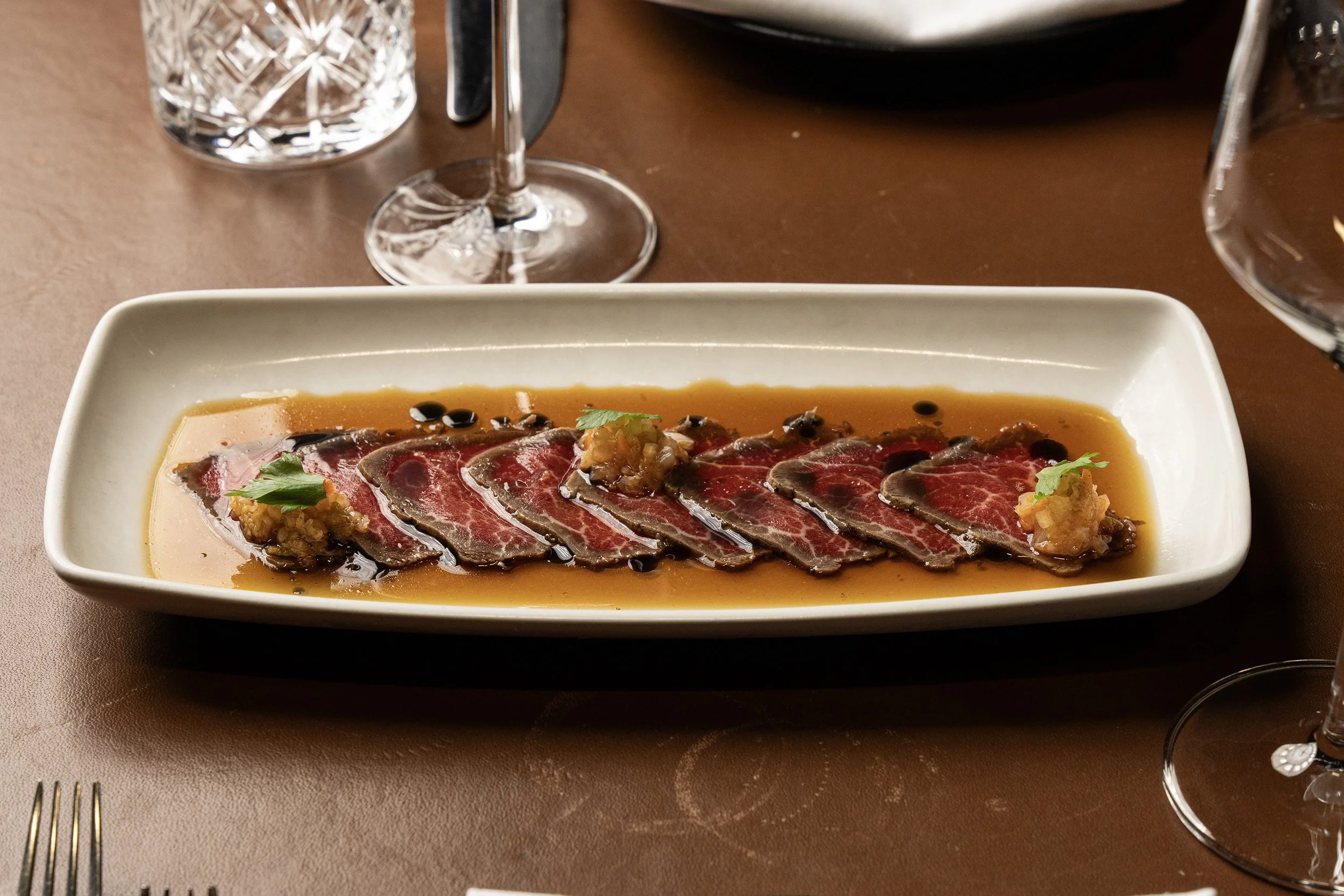 Auf einem weißen ovalen Teller sind dünne Scheiben rohes Rindfleisch mit dunkler Soße und kleinen Garnituren wie grüne Blätter, dunkle Beeren und eingelegter Ingwer. Der Tisch ist braun und es sind Wein- und Wassergläser zu sehen.