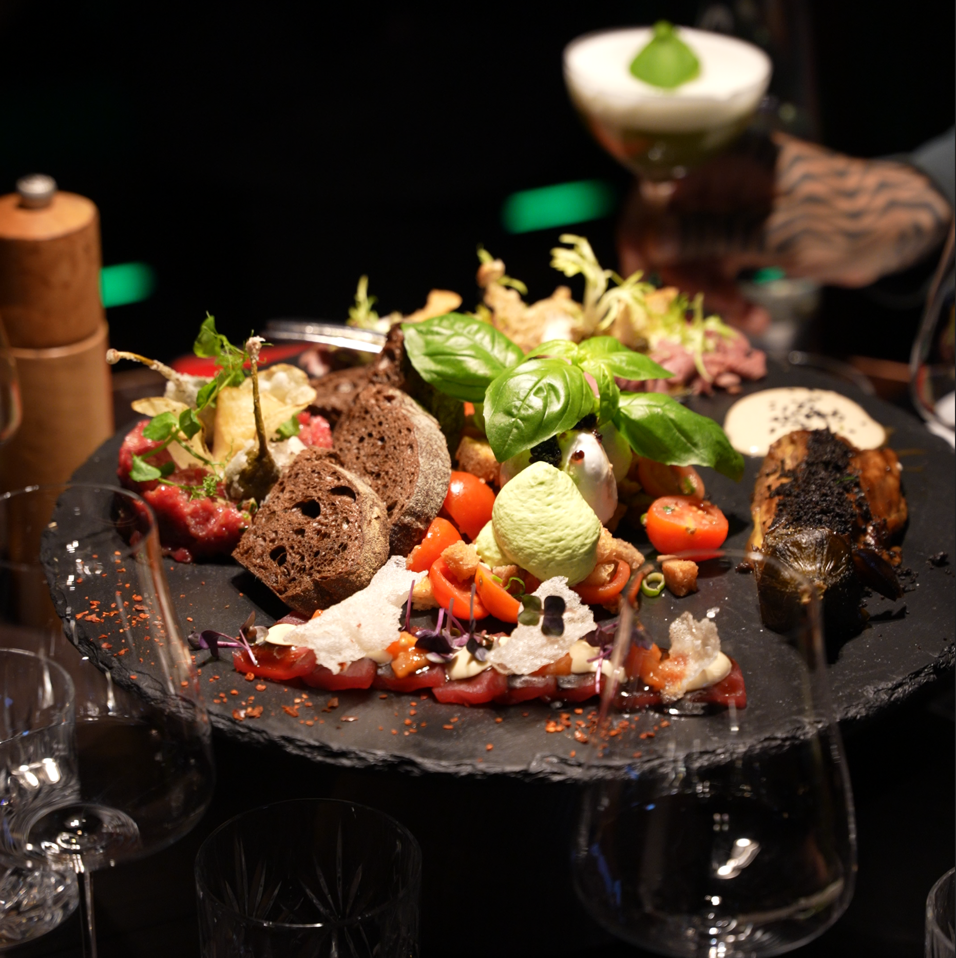 Verschiedene speisen auf einem dunklen Teller, inklusive Brot, Tomaten, frischen Kräutern, einer cremigen Sauce, Fisch und anderen Zutaten, serviert auf einem schwarzen Teller mit Gläsern im Vordergrund.