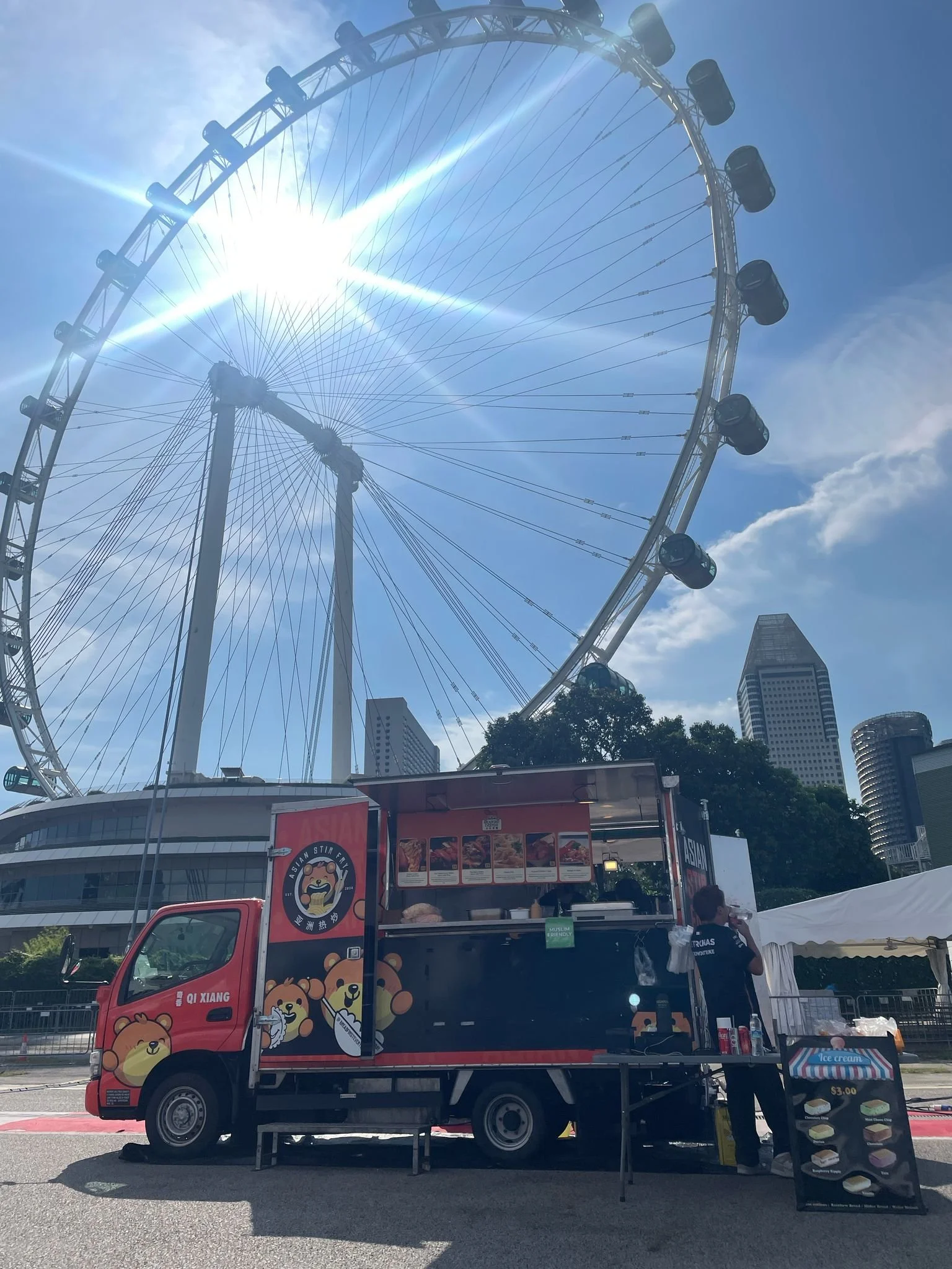 Food truck with cartoon bear design near a Ferris wheel under a sunny sky, with city buildings in the background.
