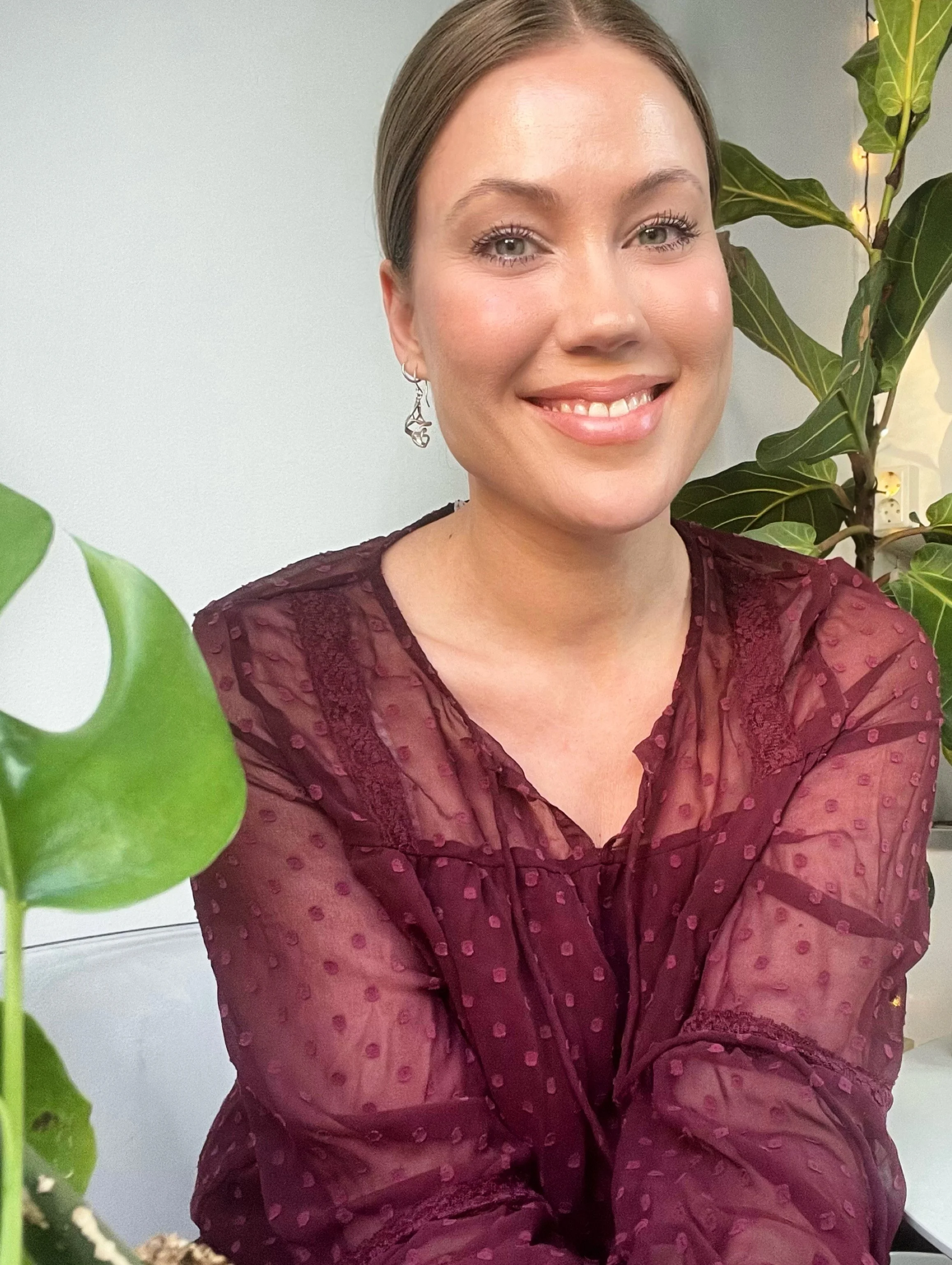 Portræt af en kvinde med kort, brunt hår, iført en burgundy farvet, gennemsigtig bluse med mønster og smykker, der sidder foran en grøn plante med lys baggrund.