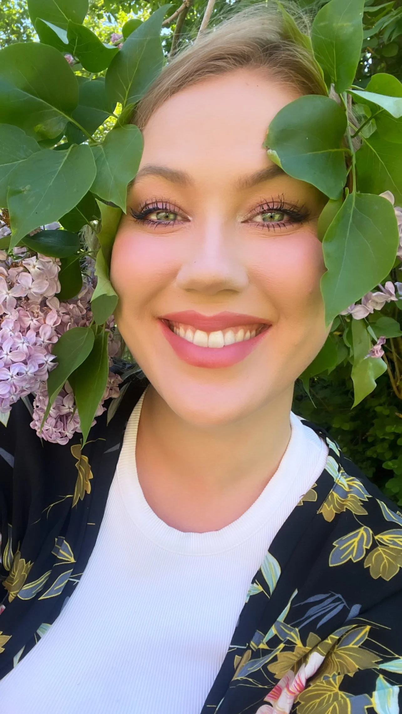 En smilende kvinde med grønne øjne, blondt hår, sminke, iført en hvid top og en sort kjole med guld- og grønne bladmønstre, omgivet af grønne blade og lyserøde blomster i baggrunden.
