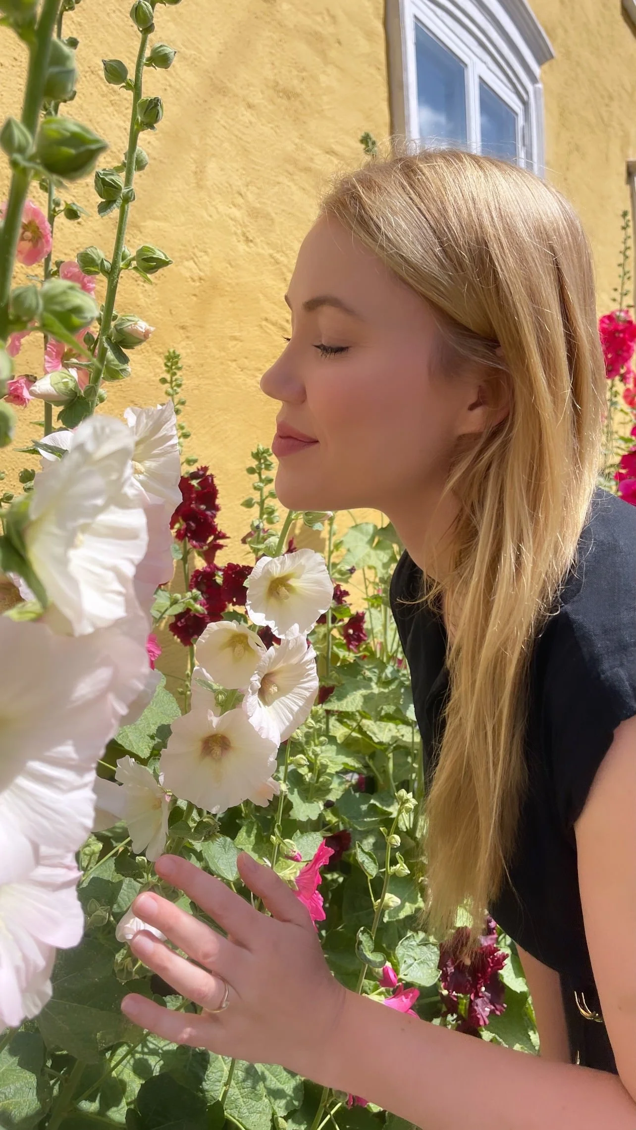 Kvinde med langt blond hår, lukkede øjne og et smil står ved en blomsterhave med lyse og mørkerøde blomster foran en gul væg og et vindue.