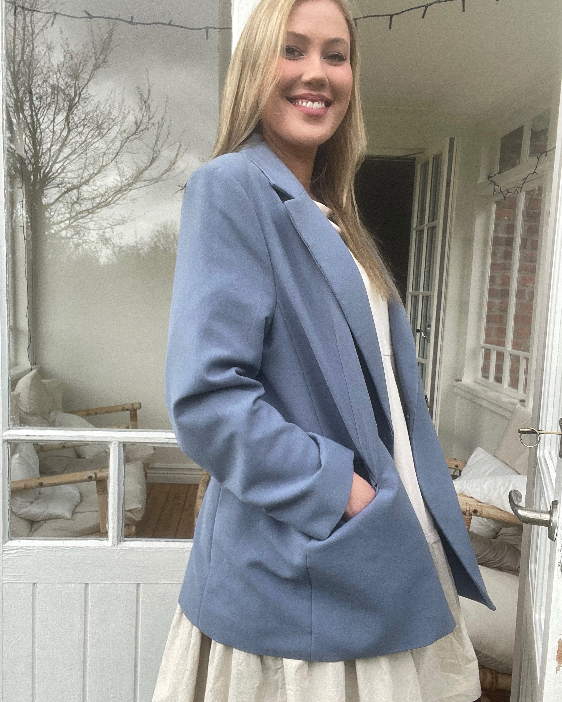 En ung kvinde smiler, iført en lyseblå blazer og en beige kjole, står foran et hvidt vindue med udsigt over et vinterlandskab med træer.