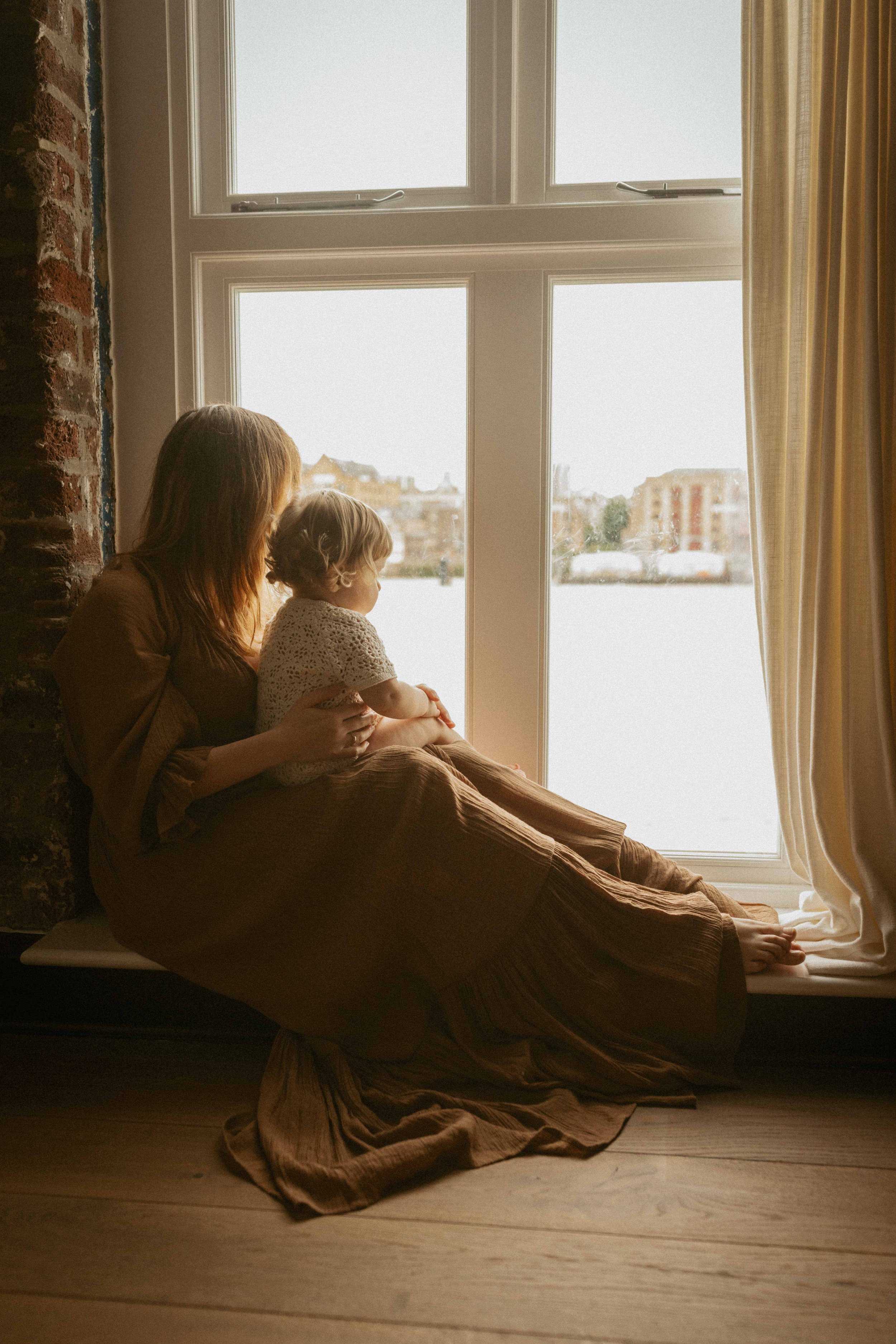A woman and a young girl sitting on a windowsill, looking outside. The woman is holding the girl. Bright daylight is visible through the large window, with buildings and a body of water outside. The room has light-colored walls, curtains, and a brick