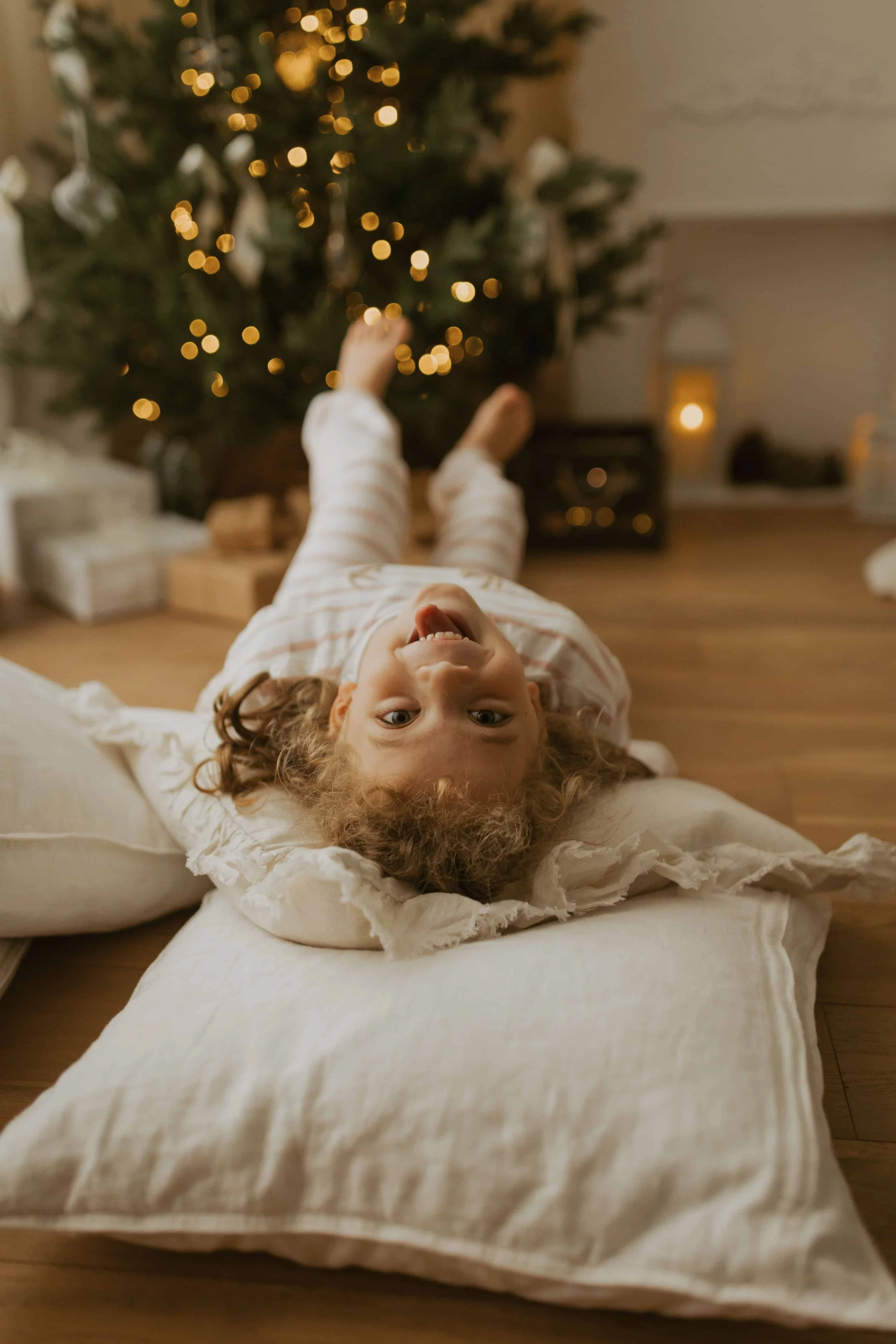 little girl in a pyjama laying on cushions by a christmas tree and fireplace with presents