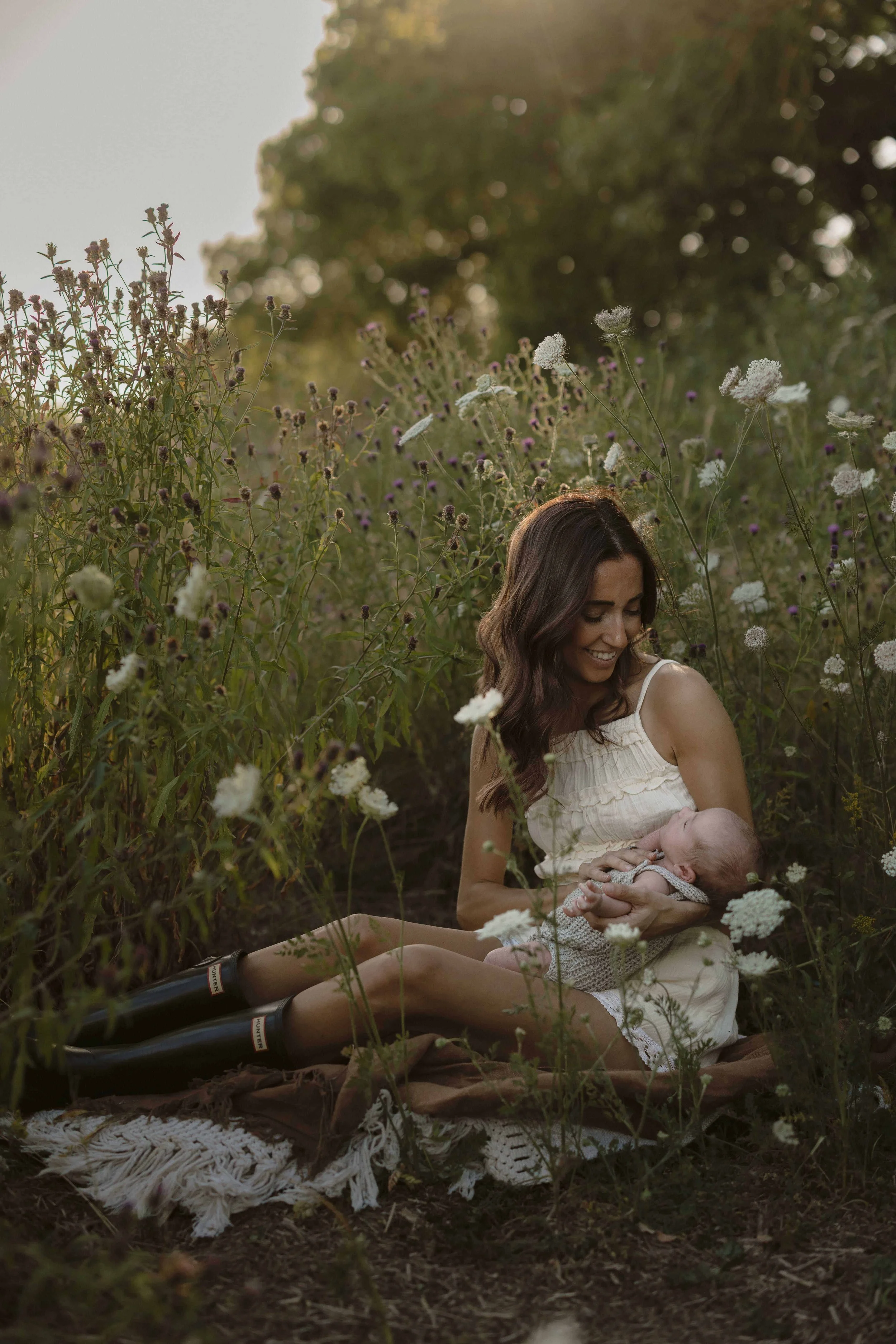 A  mum smiles as she holds a baby, sitting among wildflowers field and greenery in an outdoor newborn photography session Surrey at golden hour