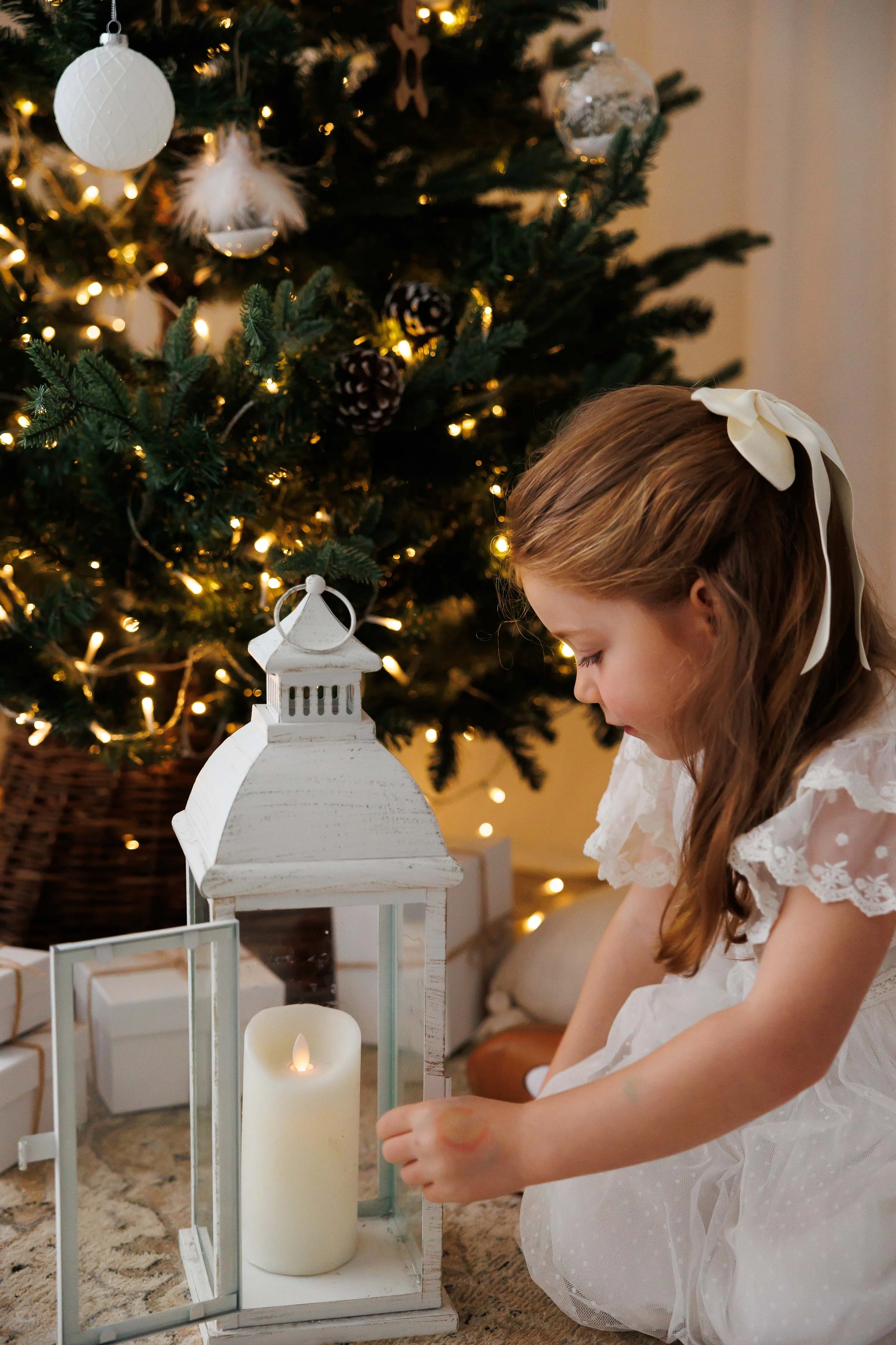 A young girl with long brown hair tied with a white ribbon, wearing a white dress with lace details, is kneeling near a Christmas tree decorated with white ornaments and warm lights. She appears to be lighting or adjusting a white lantern with a larg