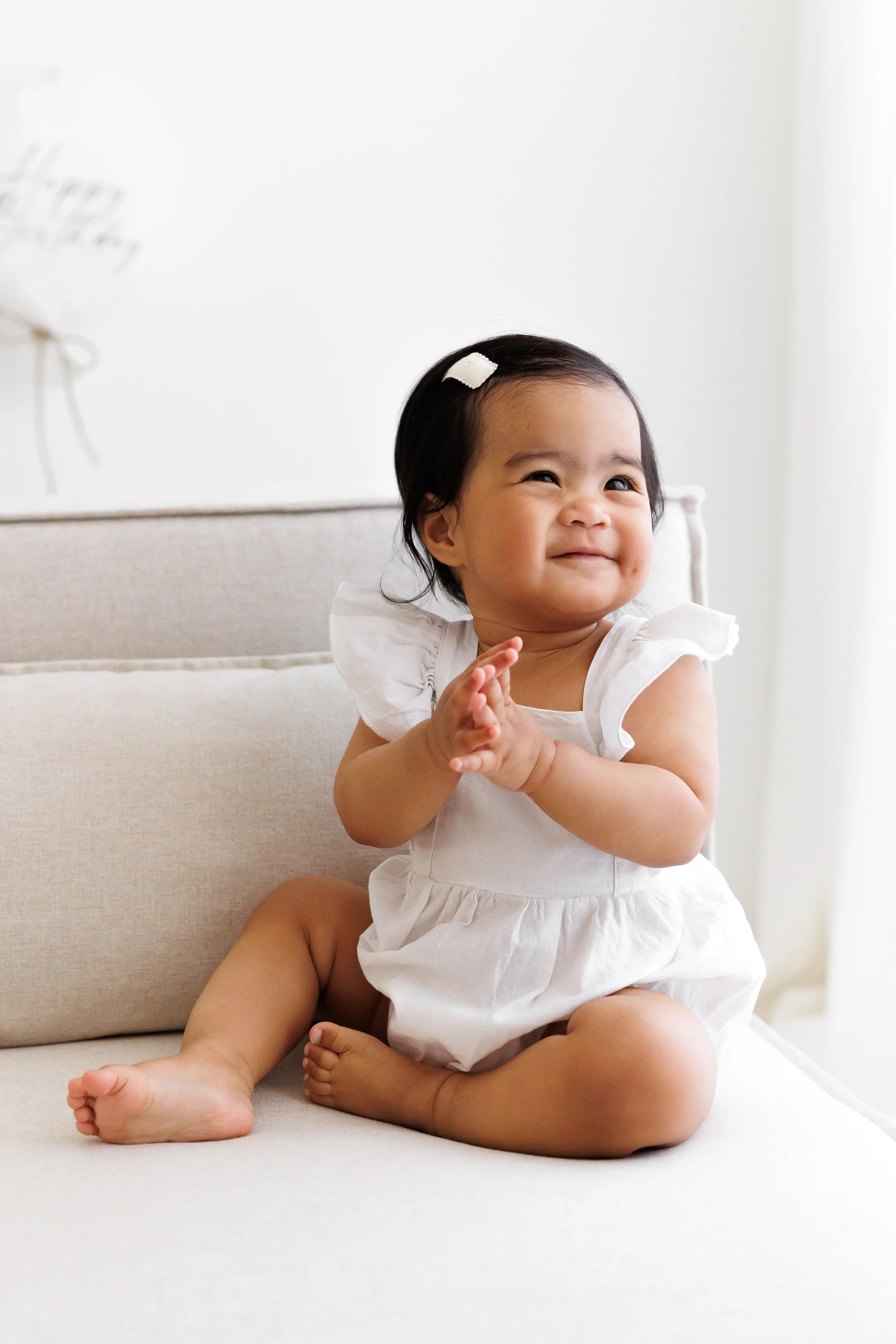 A young girl with dark hair, wearing a white dress and a hair clip, sitting on a beige sofa and smiling with her hands clasped together.