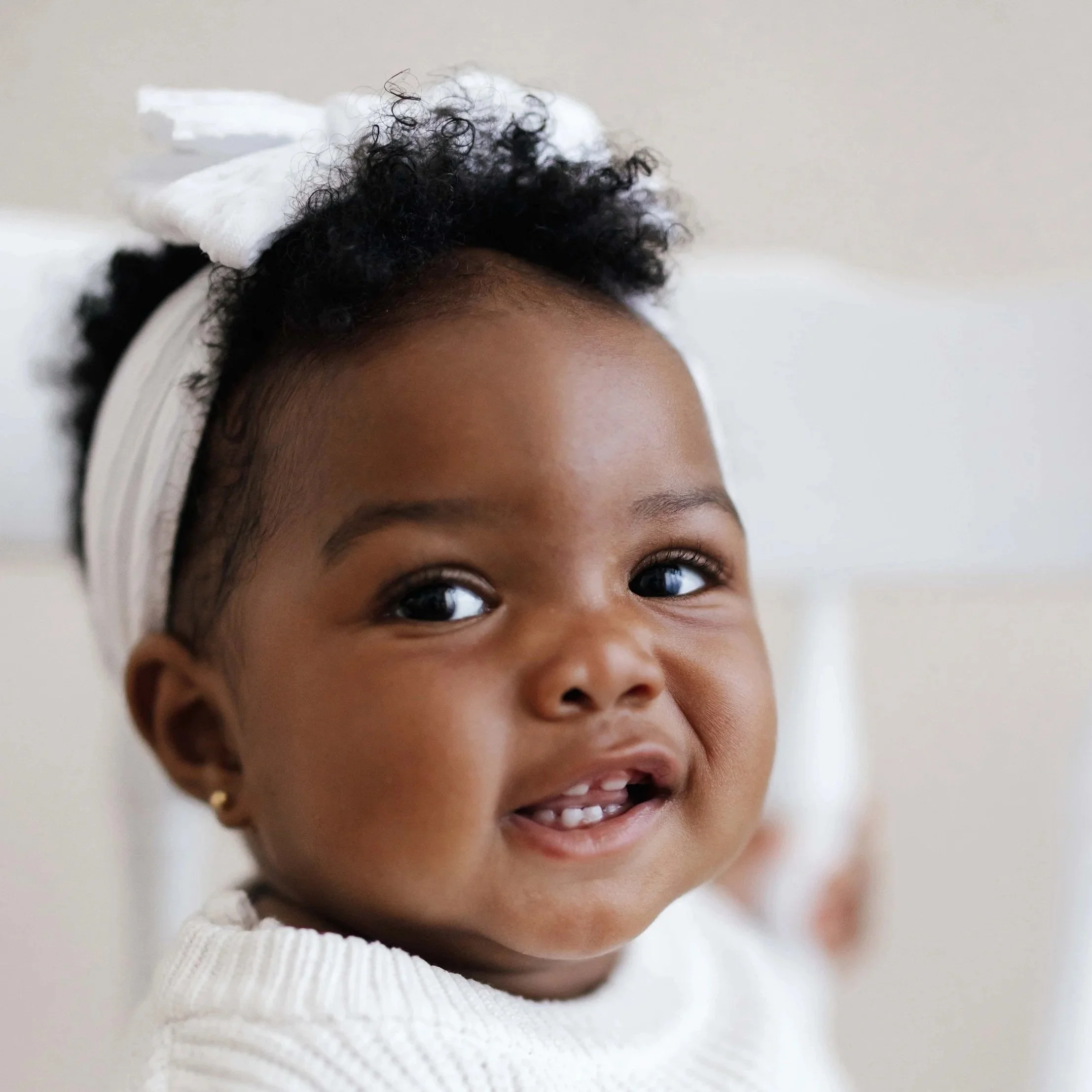 Close-up of a smiling baby girl with curly black hair wearing a white headband and white sweater.