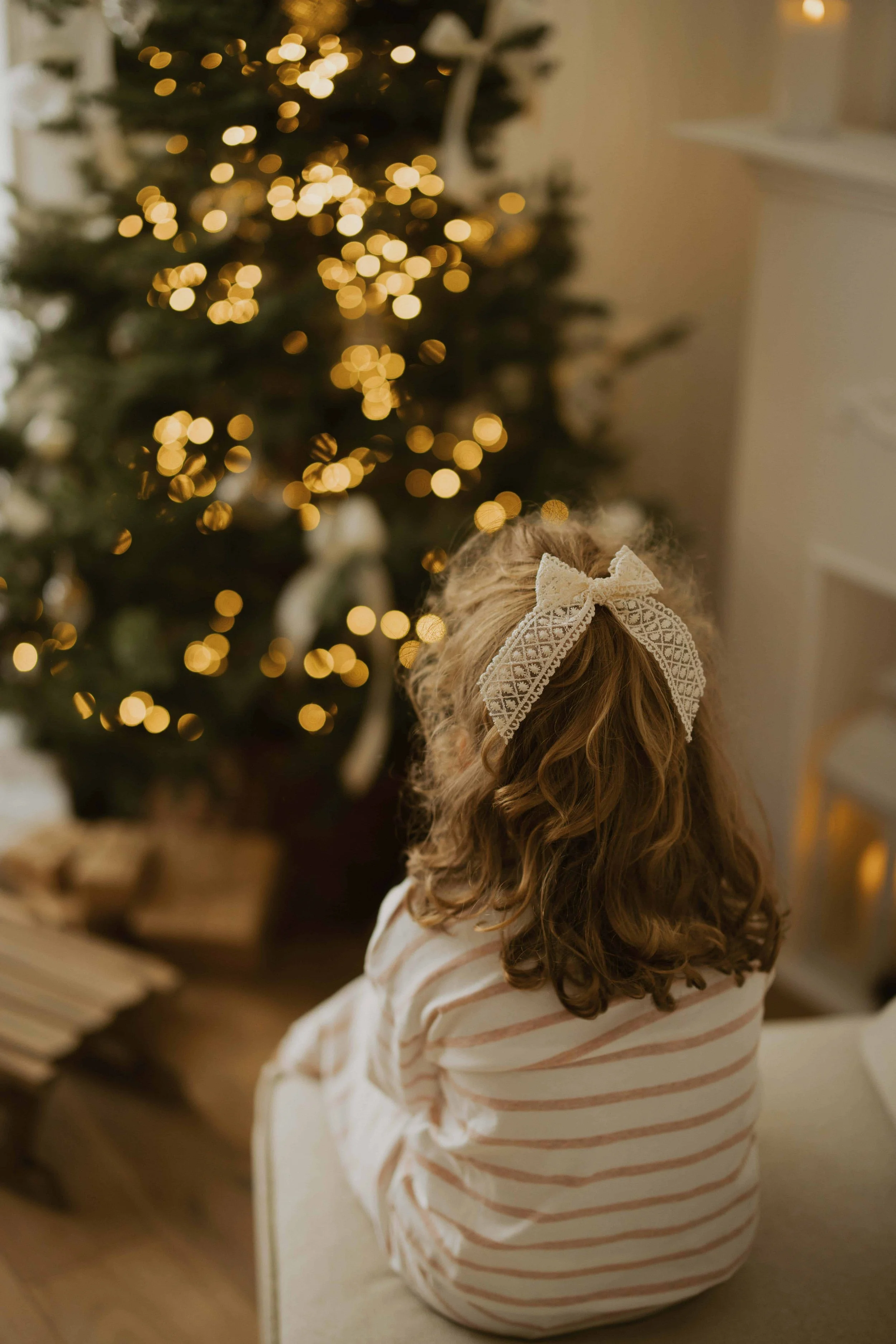 A young girl with curly hair wearing a lace headband and striped pajamas, sitting on the floor in front of a decorated Christmas tree with blurred lights.