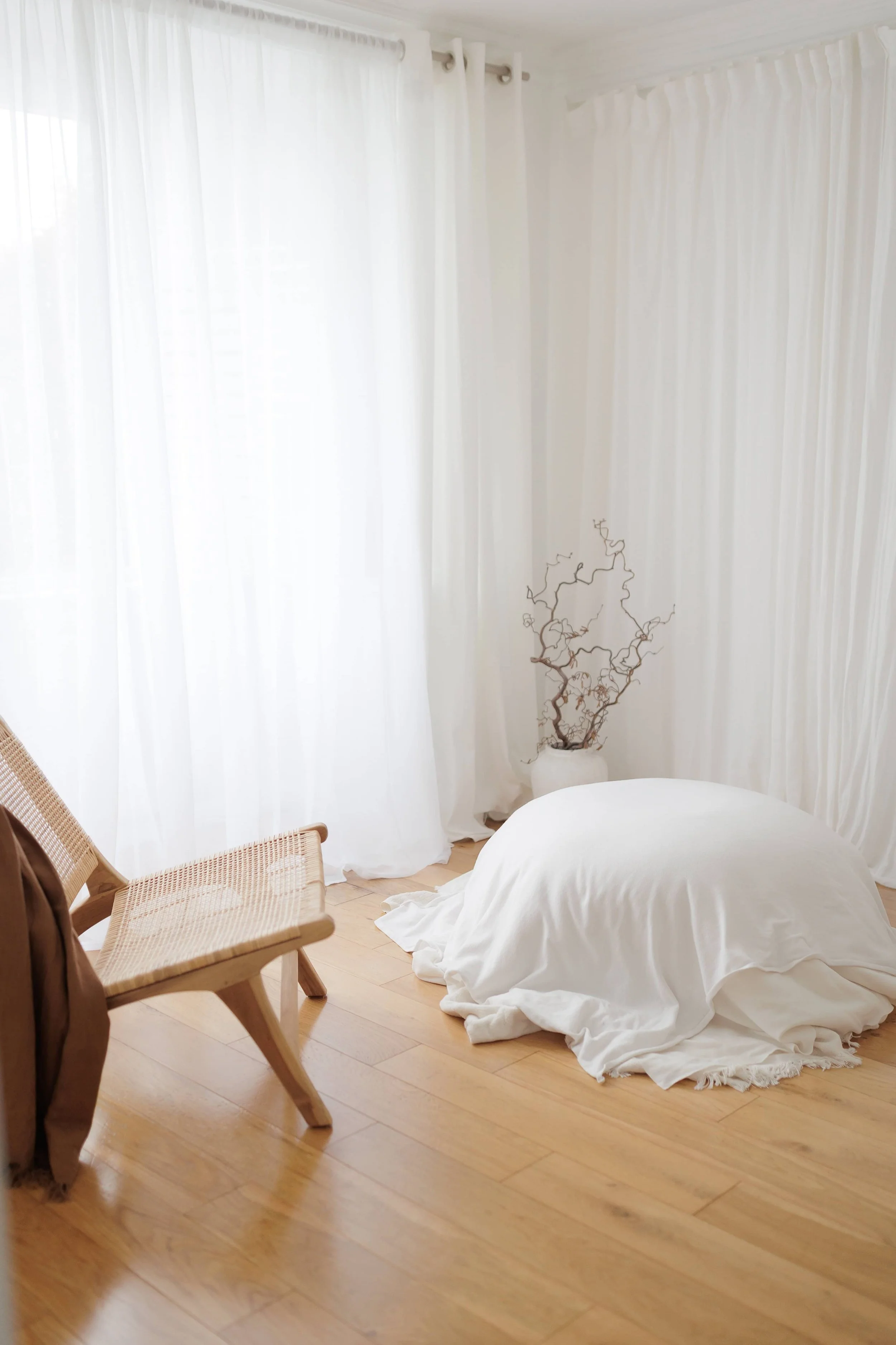 A room with white curtains, a wicker chair, a brown jacket on the chair, a white cloth-covered object on the floor, and a white vase with curly twigs in the corner.