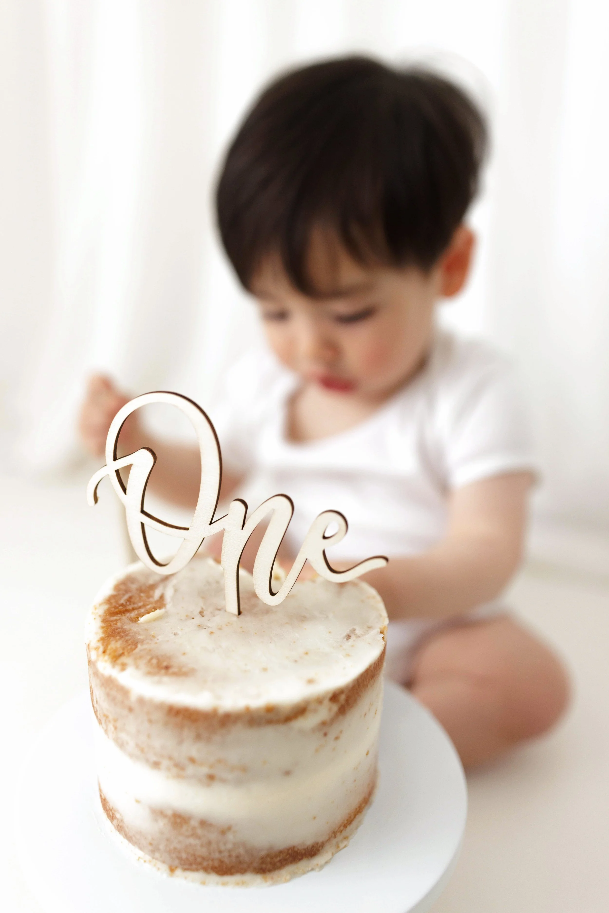 Close-up of a small layered birthday cake with white frosting, decorated with a wooden "One" cake topper, placed on a white stand.