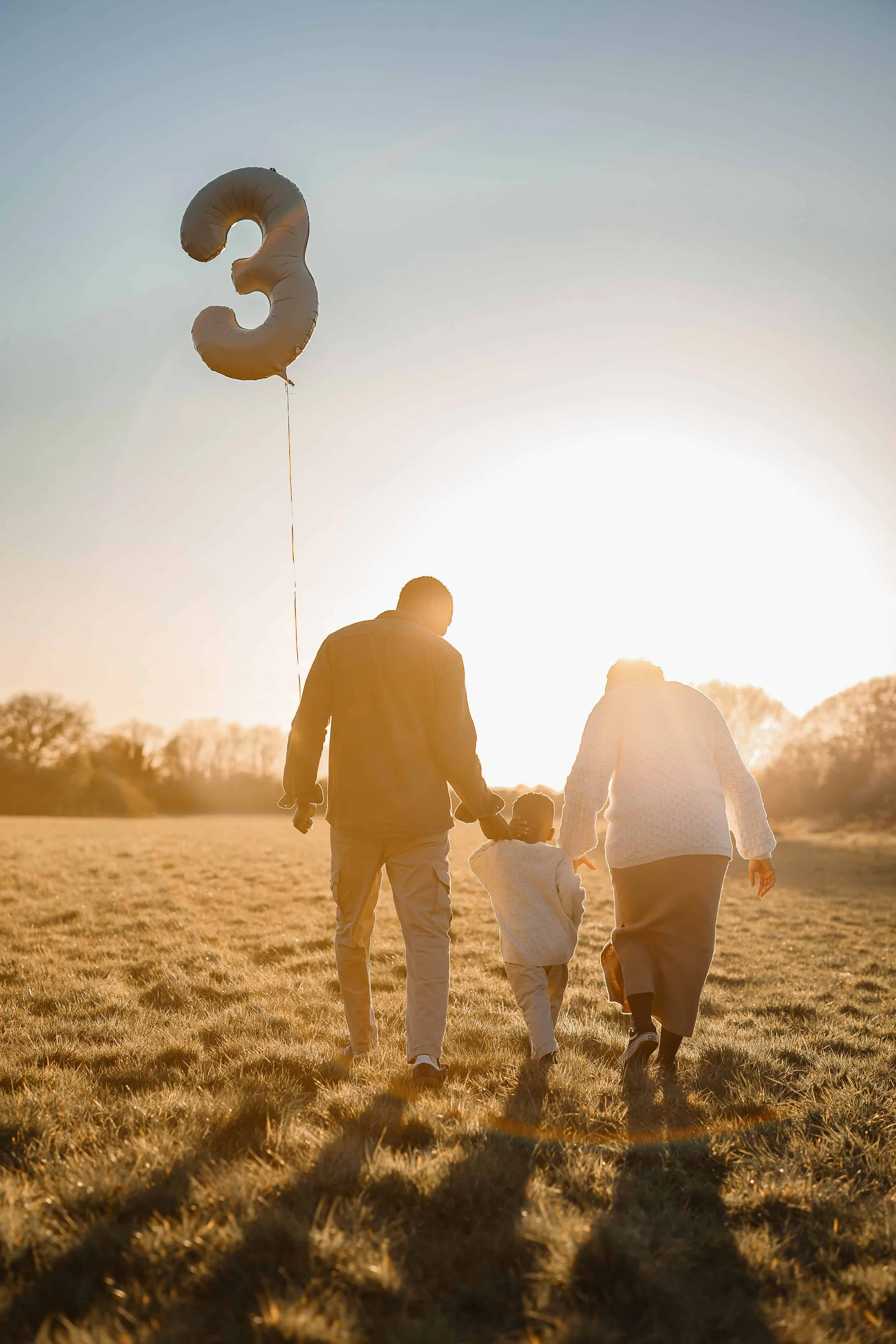 A family of three walking hand in hand in a grassy field at sunset with a large balloon shaped like the number three in the sky.