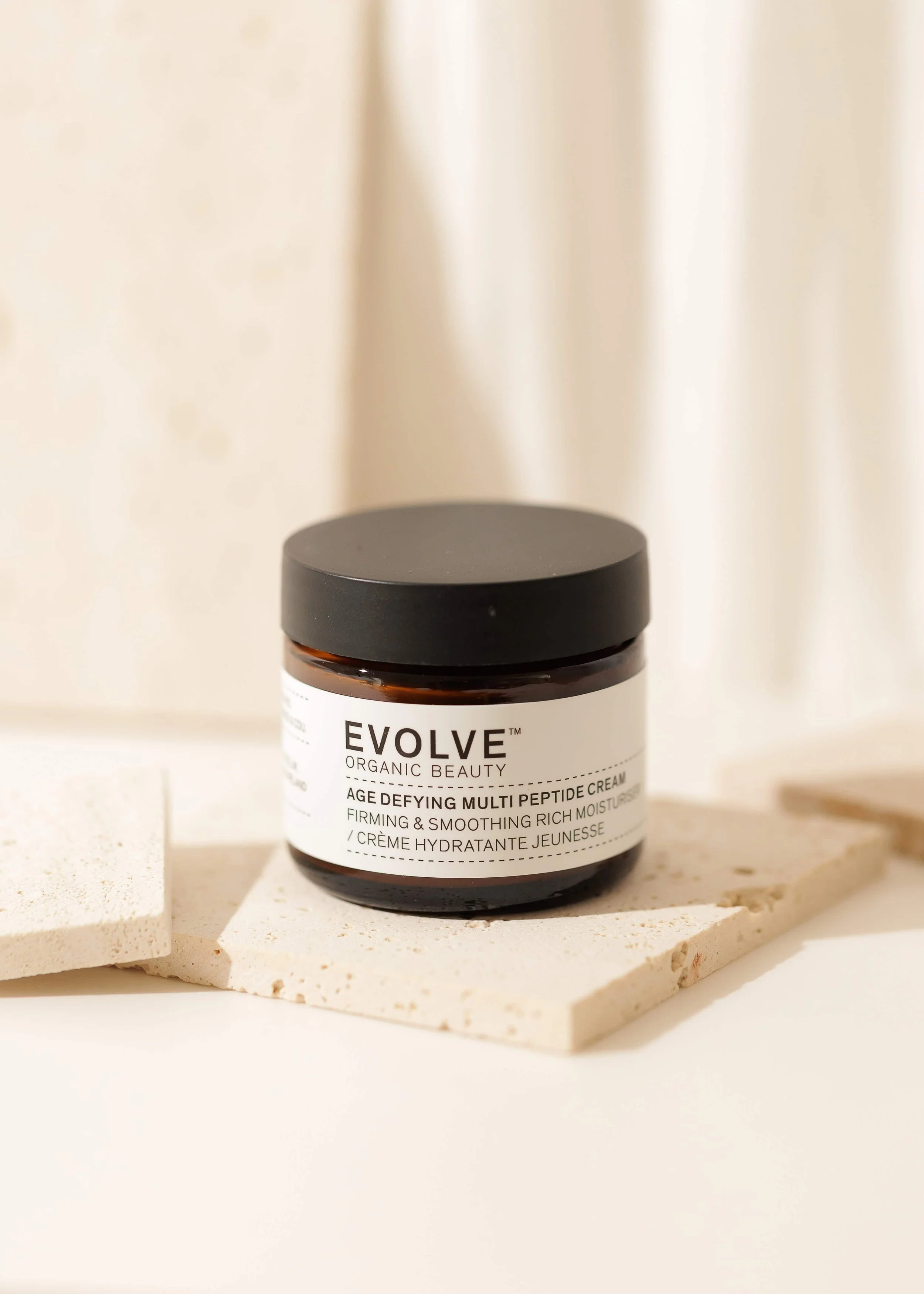 Bottle of EVOLVE Organic Beauty Age Defying Multi Peptide Cream on a white surface with stone tiles in the background.
