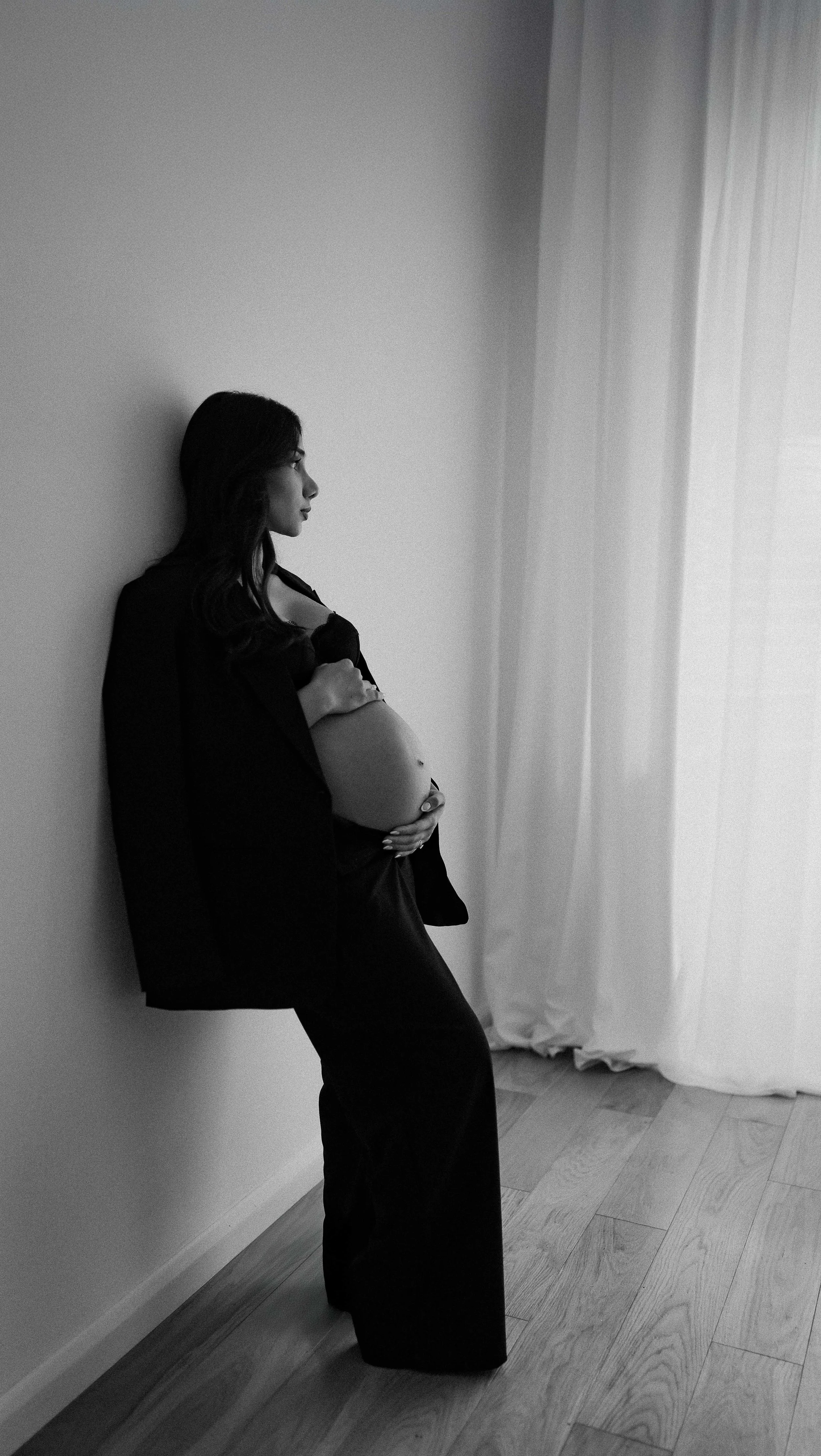 Black and white photo of a pregnant woman in black clothing, standing against a wall, with her hand on her belly, looking thoughtfully out of the window.