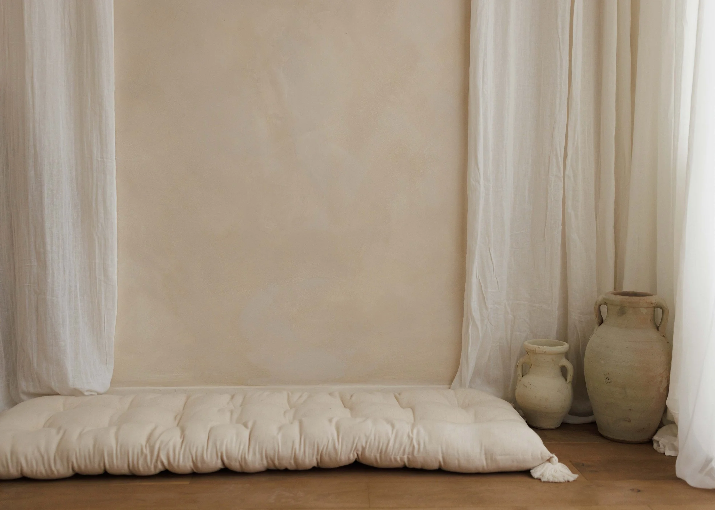 A minimalist in-home studio with cream-colored curtains, a beige wall, and two large antique-style ceramic vases on the floor, with a large white quilted cushion or bed in the foreground.