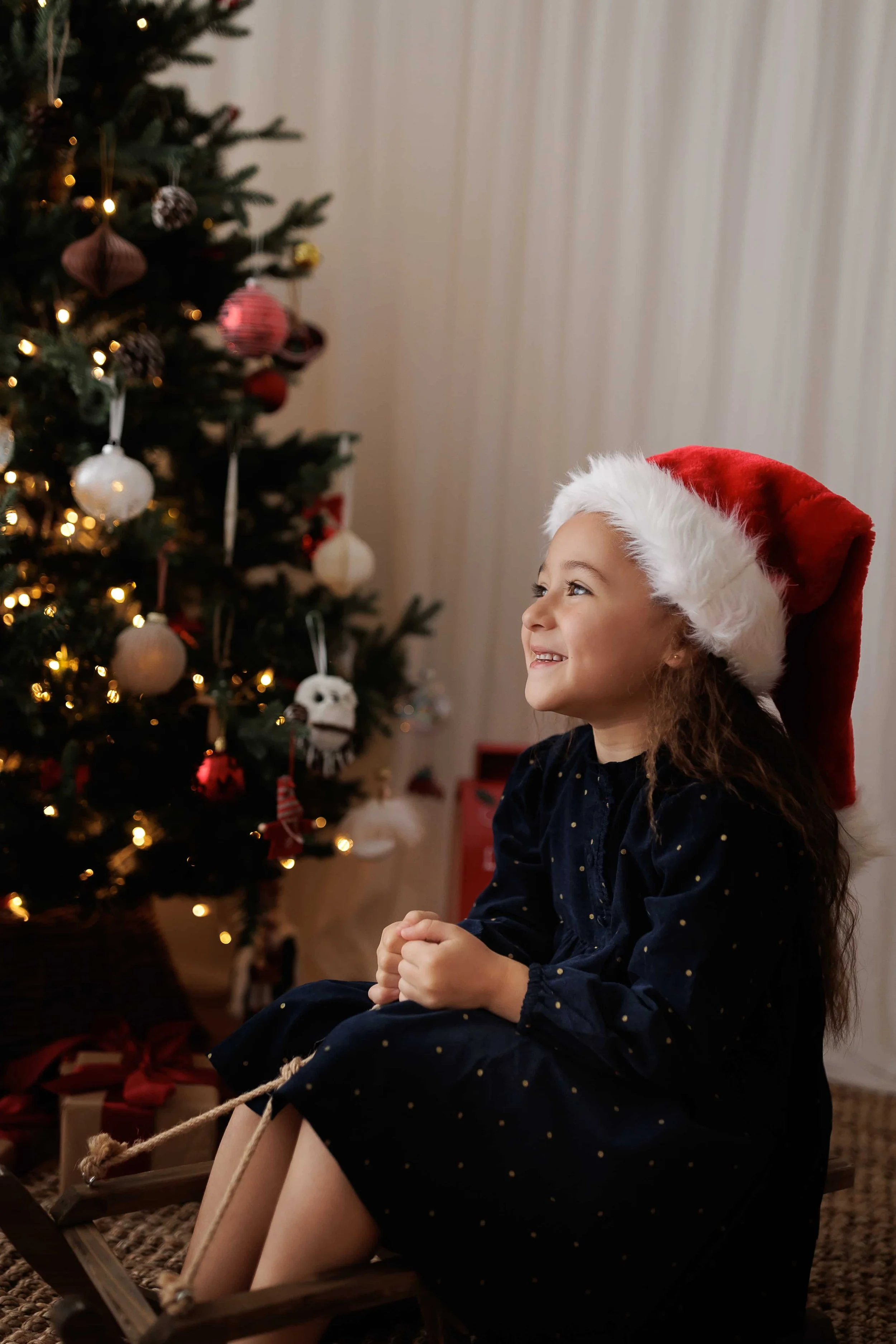 A young girl wearing a Santa hat, sitting on a wooden sled near a decorated Christmas tree with ornaments and lights, smiling and looking cheerful.