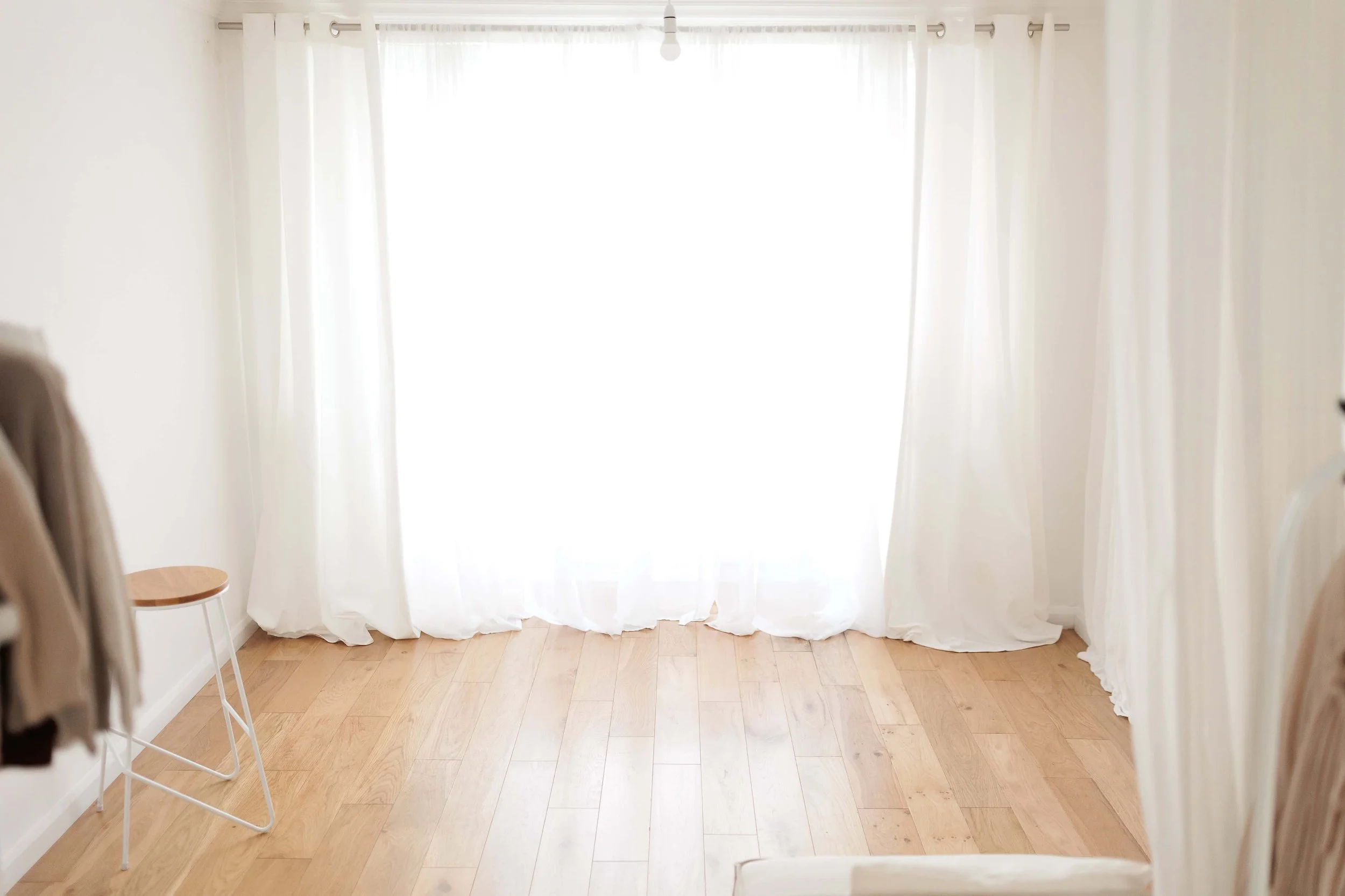 Bright, empty room with white curtains and wooden flooring, with a small wooden stool on the left and a portion of a bed on the right.