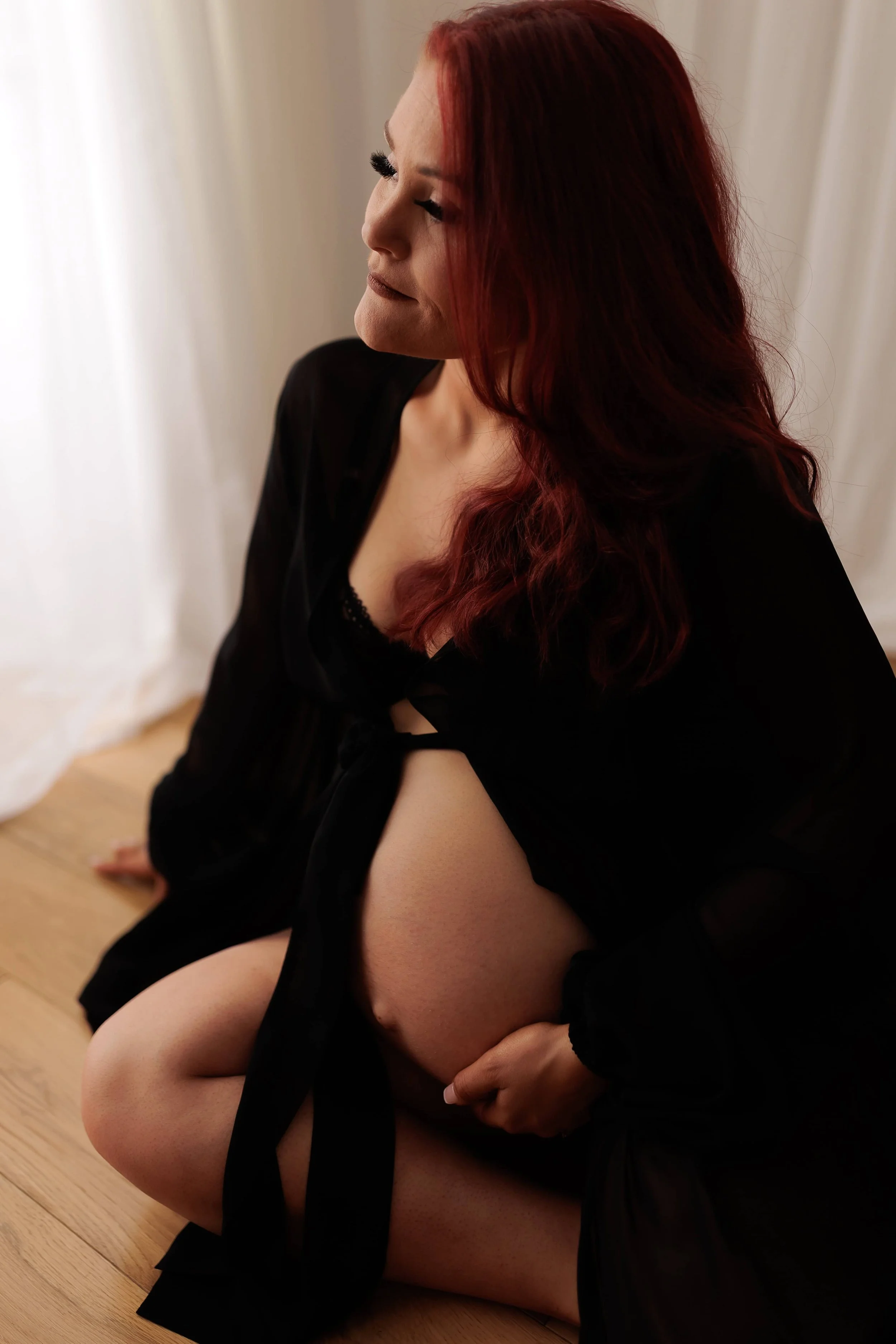 A woman with long, red hair, wearing a black outfit, sitting on a wooden floor, with her eyes closed and a serene expression.