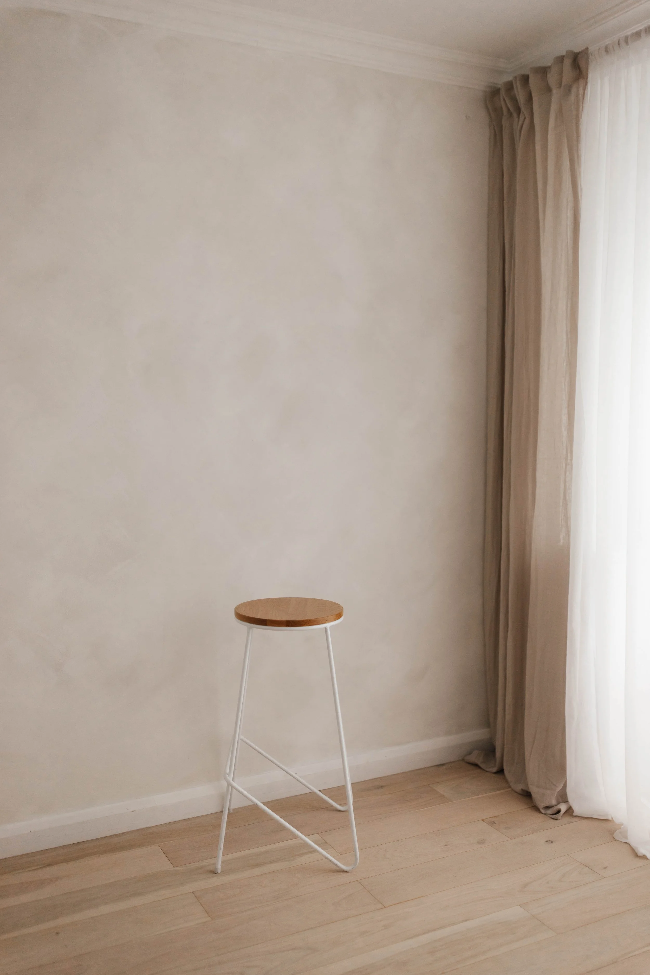 A minimalist photography studio in Surrey with a white wall, beige curtains, light wooden floor, and a small stool with a wooden seat and white metal legs.