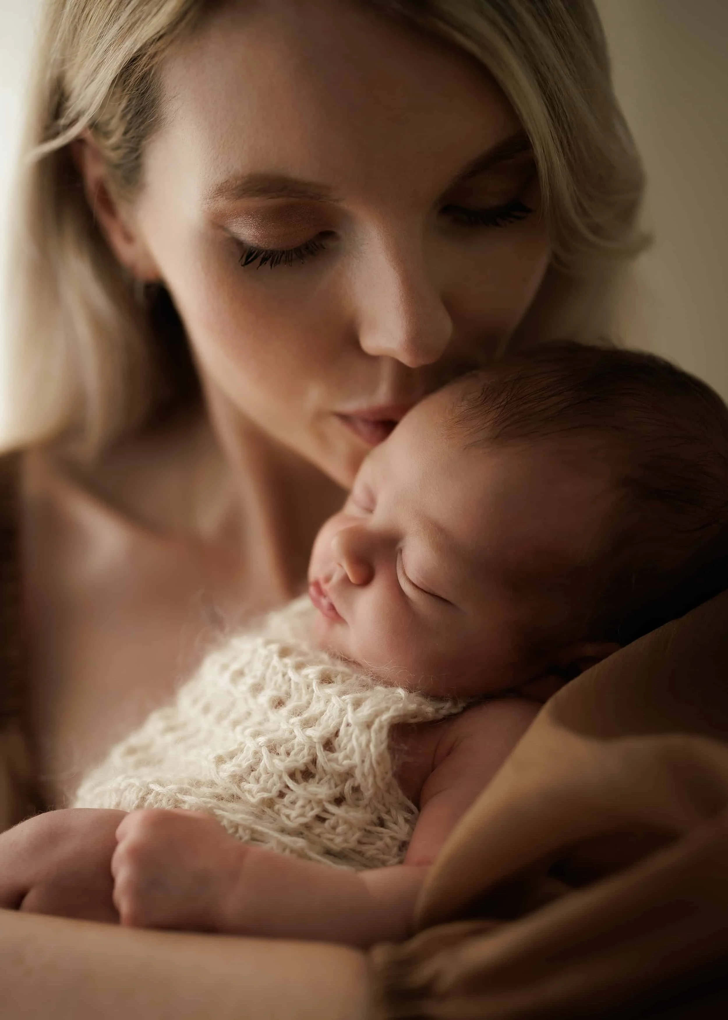 A close-up of a woman with blonde hair gently holding a sleeping baby, both with their eyes closed, in a warm and tender moment.