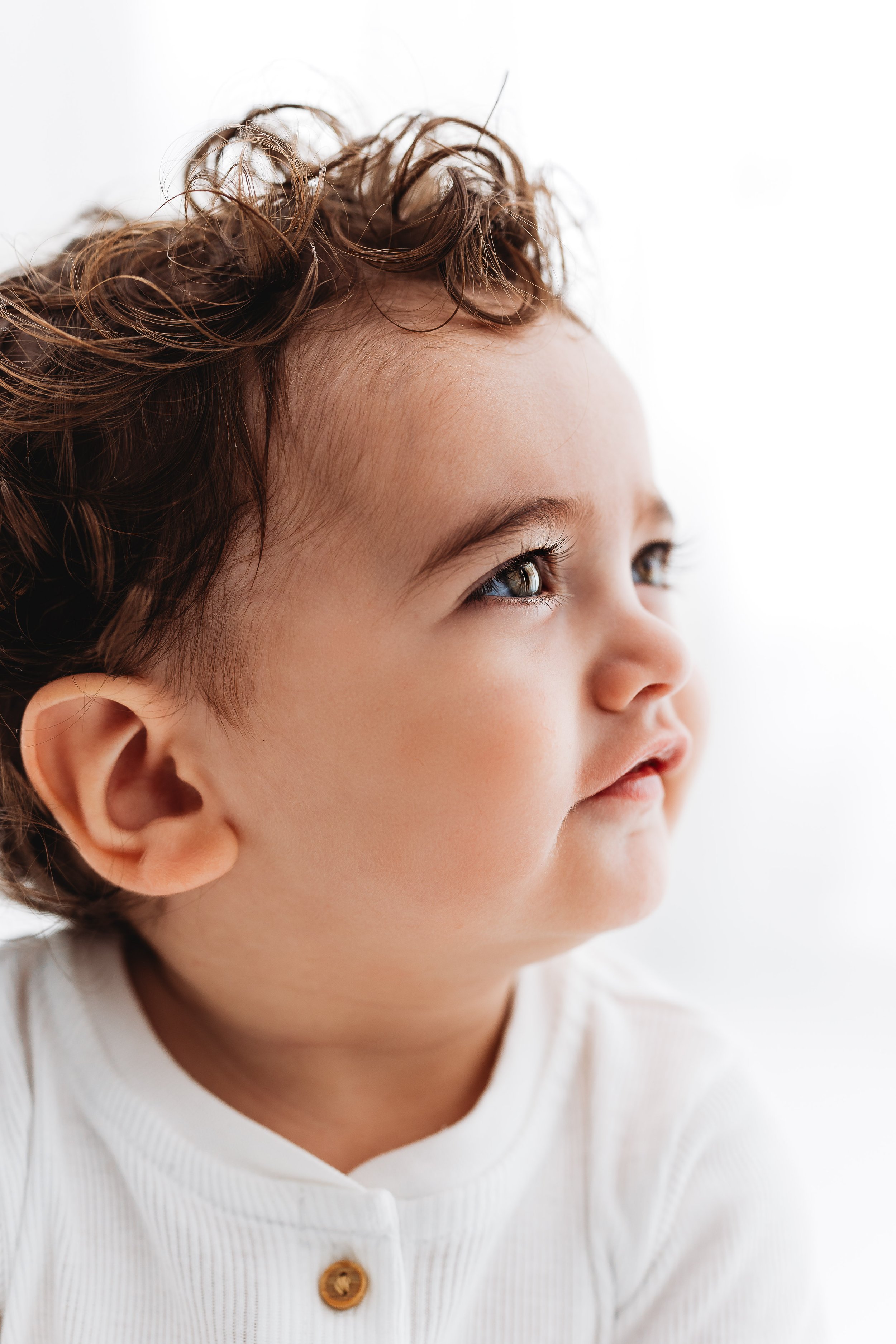Close-up of a baby with curly hair looking to the side, wearing a white shirt.