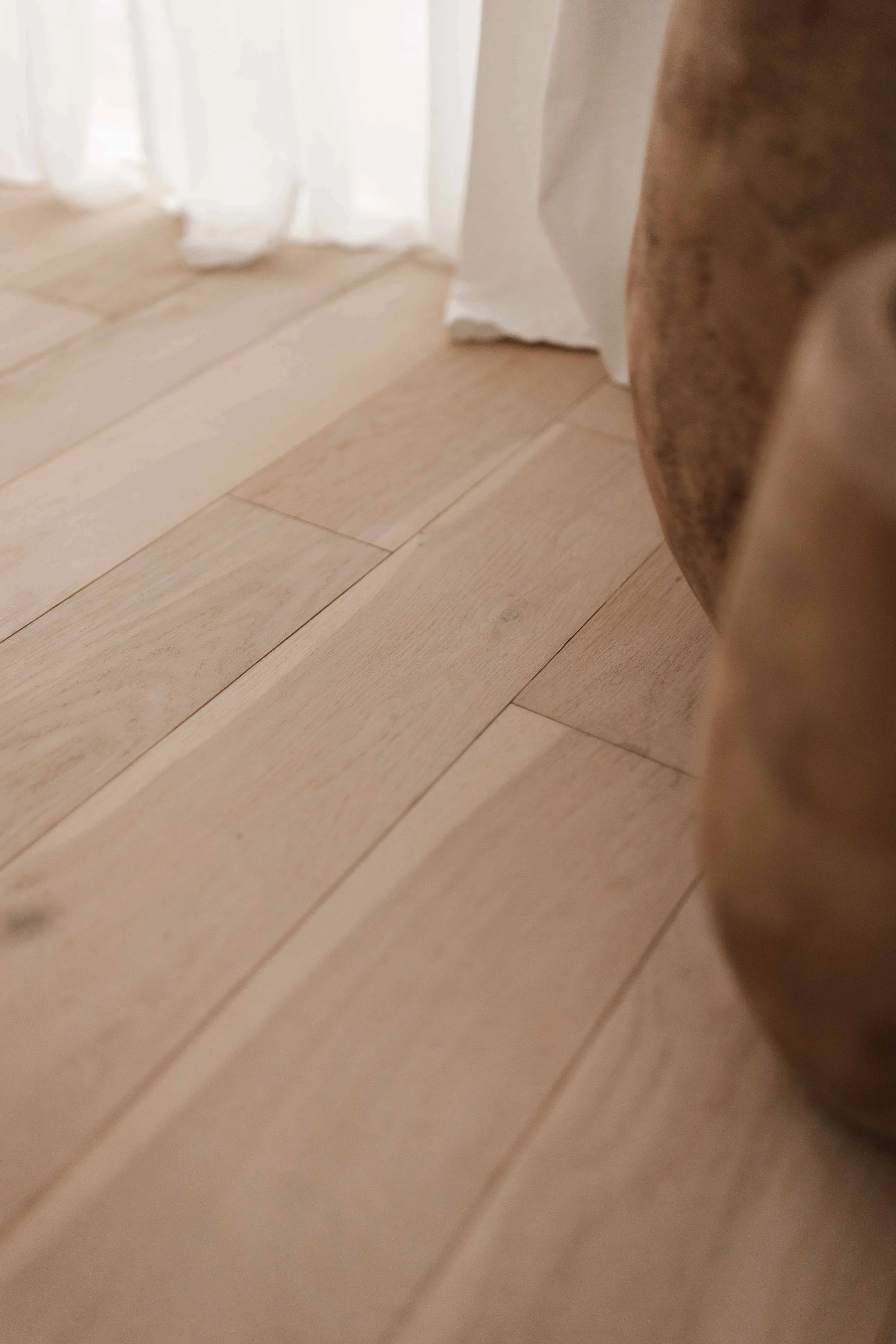 Close-up of a light wooden floor next to a white curtain and beige furniture visible on the right side, natural light studio photography in Surrey