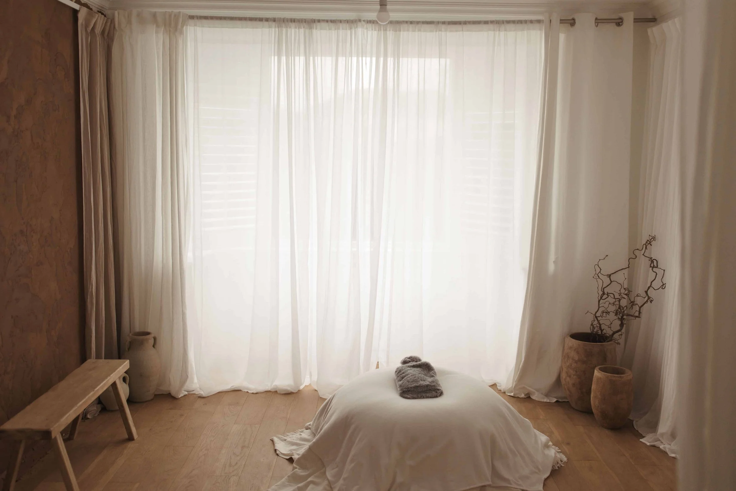 A cozy bedroom with natural light coming through a large window covered with sheer white curtains. The room features neutral tones, with a bed draped in white bedding, a plush gray pillow, and decorative vases and a potted plant on the sides.