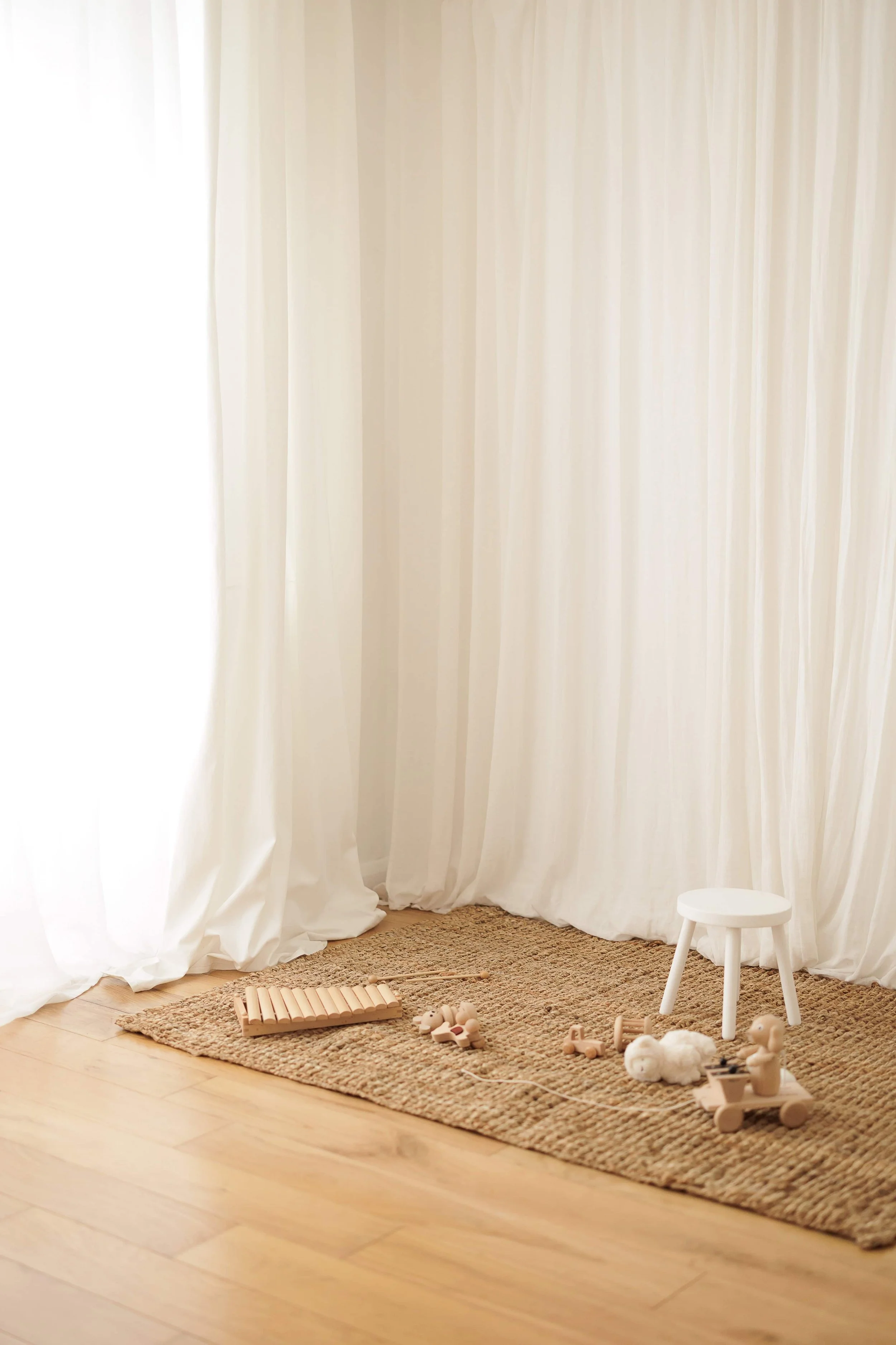A beige rug on a wooden floor with white curtains in the background. Wooden toys, a small white stool, and a xylophone are arranged on the rug.