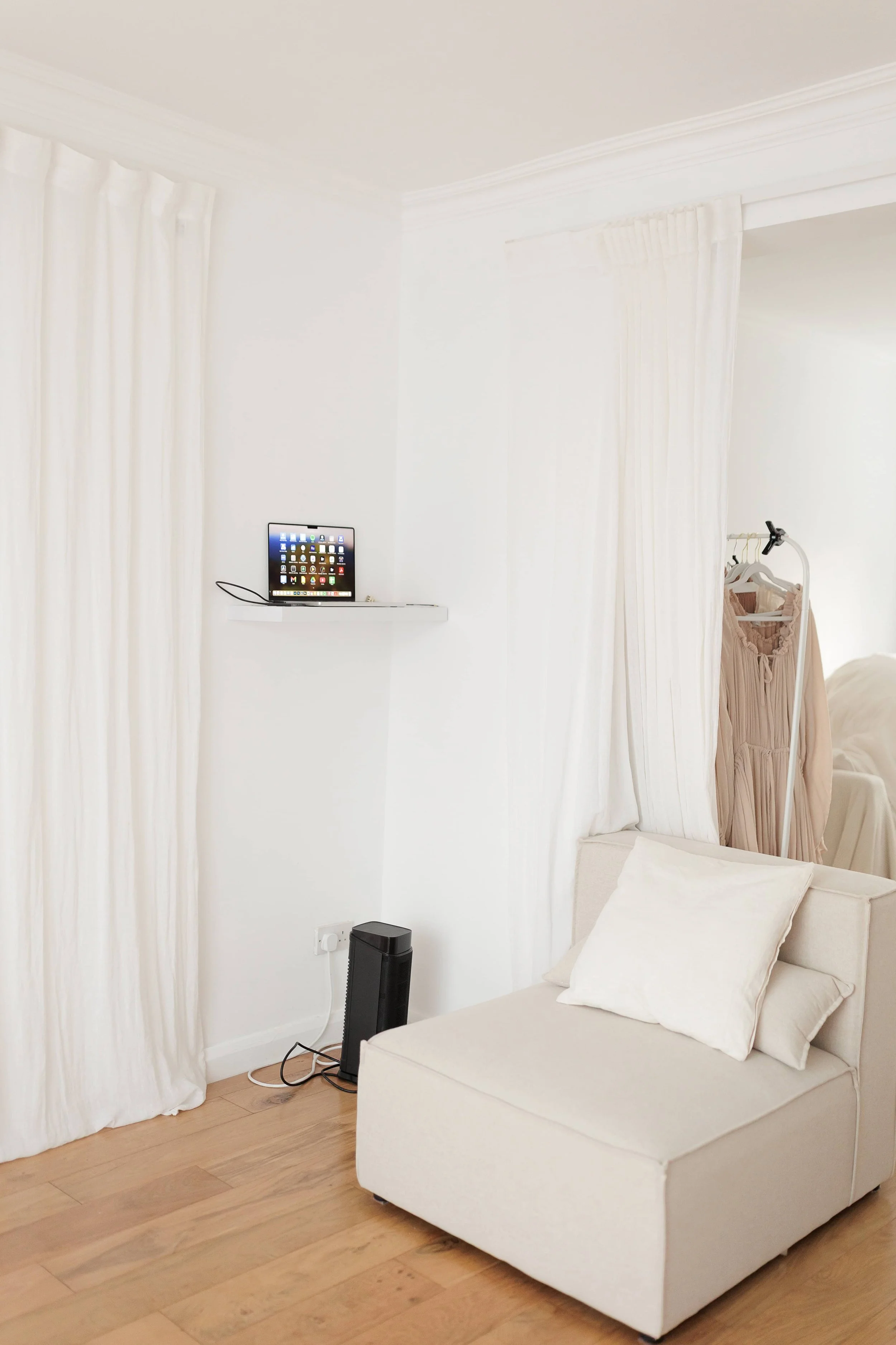 A cozy corner of a room with white curtains, a small white shelf with a tablet, a white armchair with pillows, a black tower fan, and an adjacent space with a clothing rack and hanging dress.