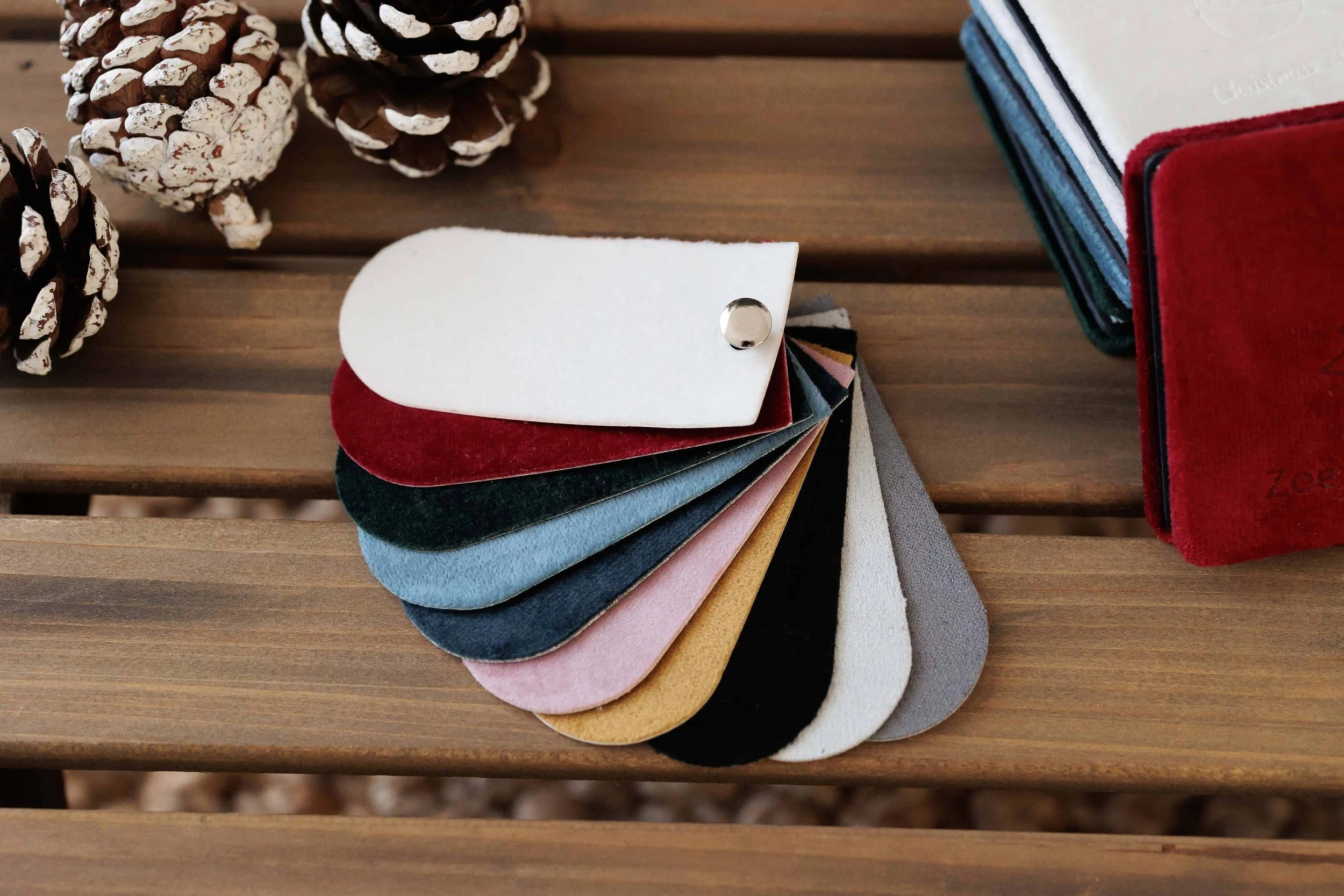 A color palette with various fabric swatches in different colors, secured with a snap button, on a wooden table surrounding pine cones and some fabric samples.