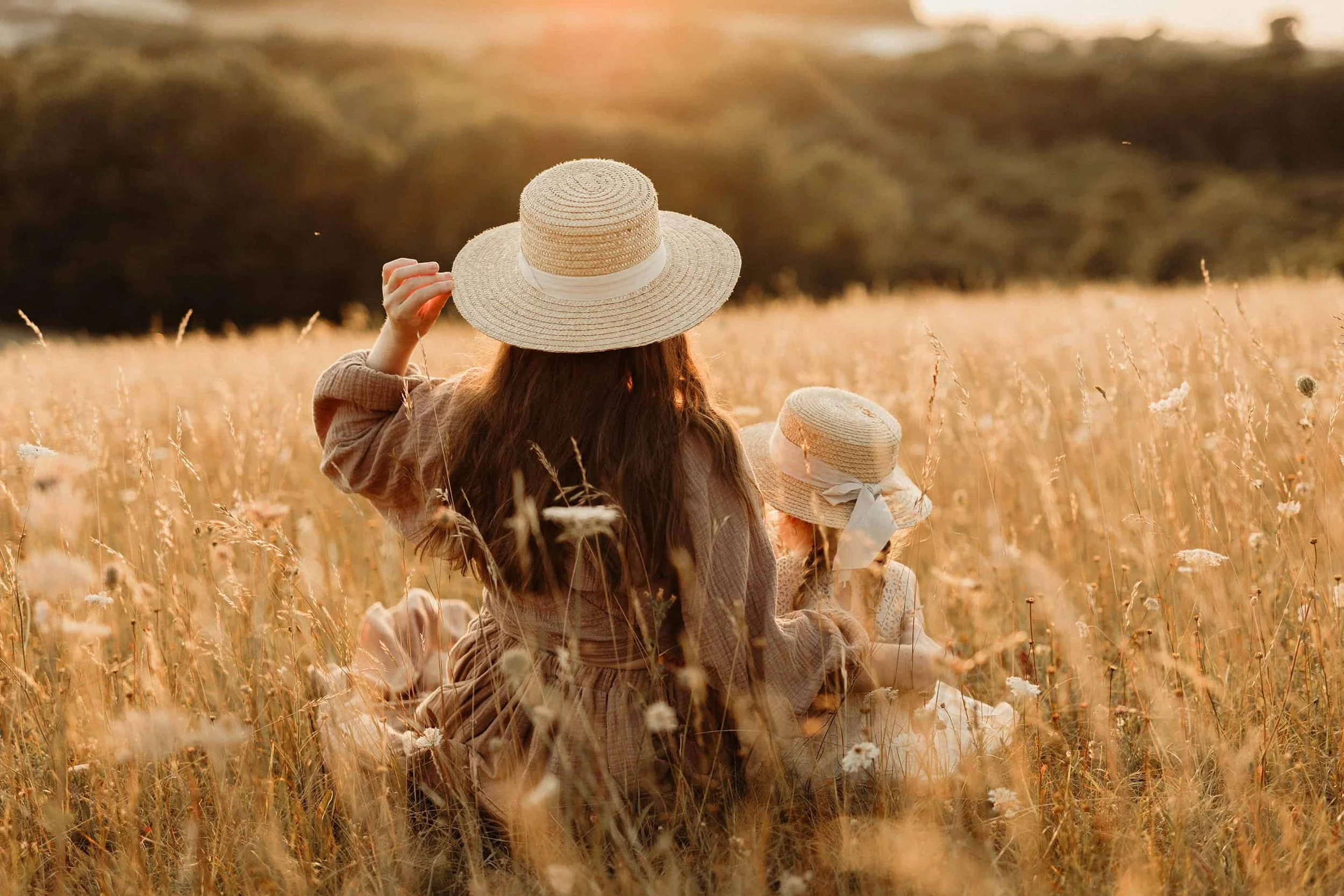 A woman and child sitting in a wheat field during sunset, both wearing wide-brimmed straw hats, facing away from the camera.