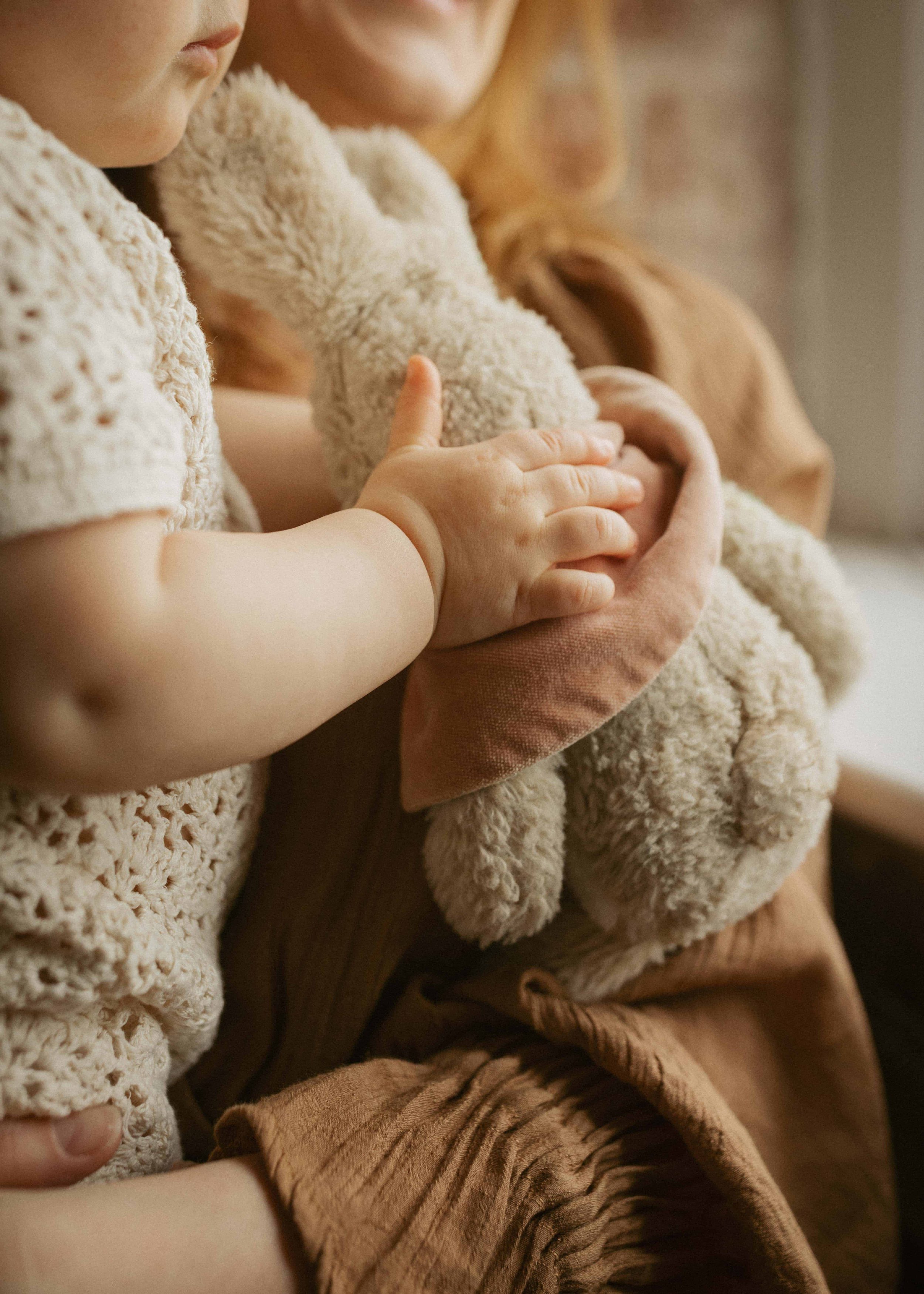 Close-up of a woman and child holding a plush teddy bear, both wearing cozy, textured beige sweaters, with their faces out of view.