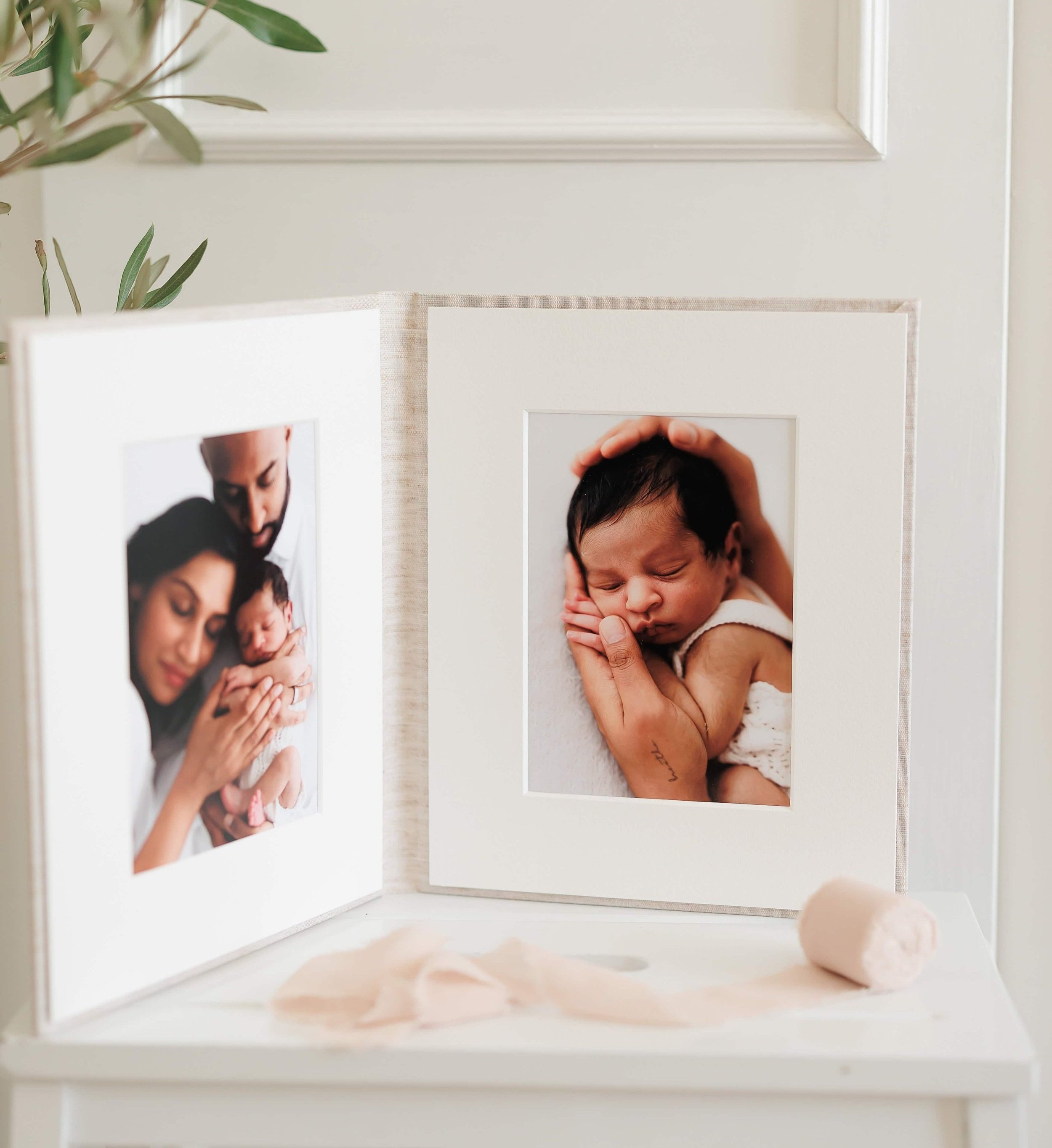 Photographs of a newborn baby displayed in a folding frame on a table, with greenery in the background. One photo shows the baby being held by parents, and another shows the baby sleeping with hands gently cradling their head.