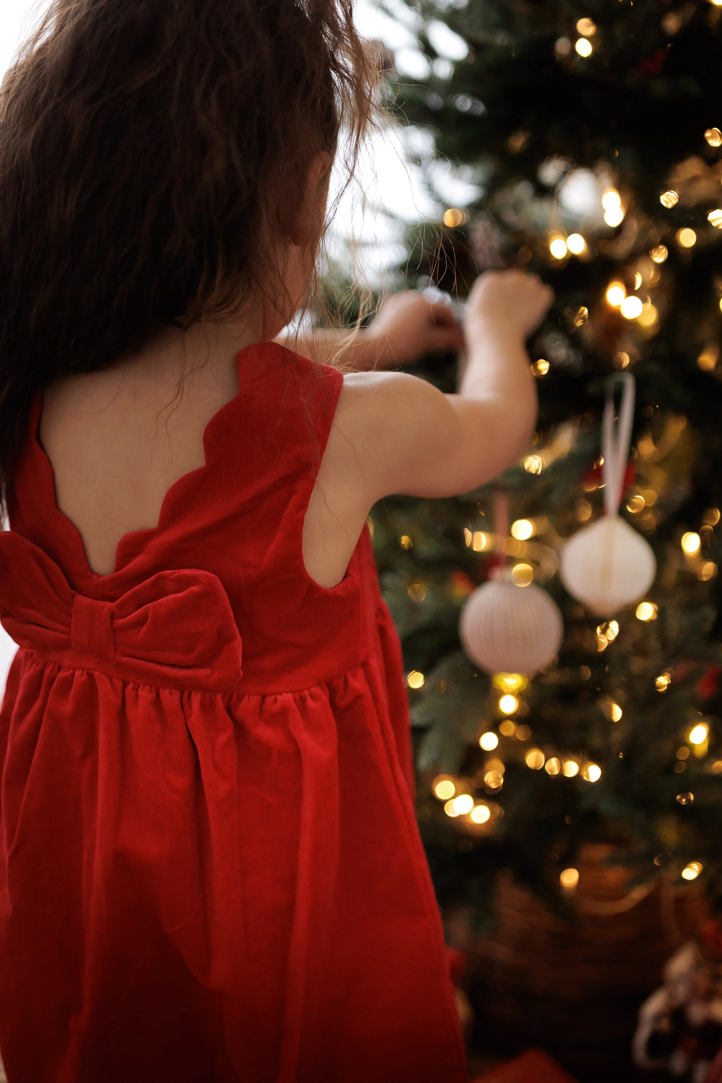 A young girl in a red dress decorating a Christmas tree with white ornaments.