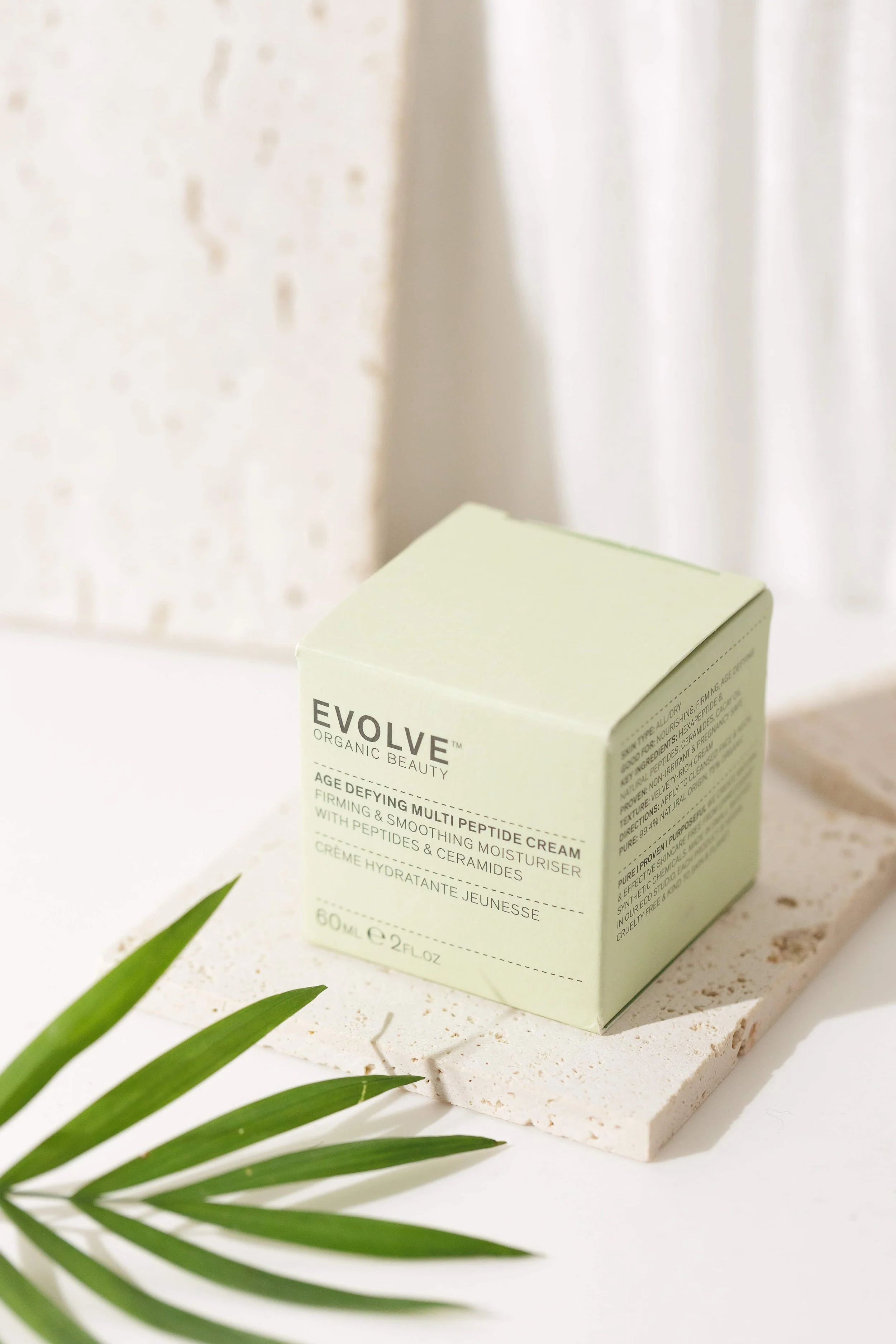 A light green box of EVOLVE organic beauty age defying multi peptide cream on a white textured surface, with a green leaf in the foreground.