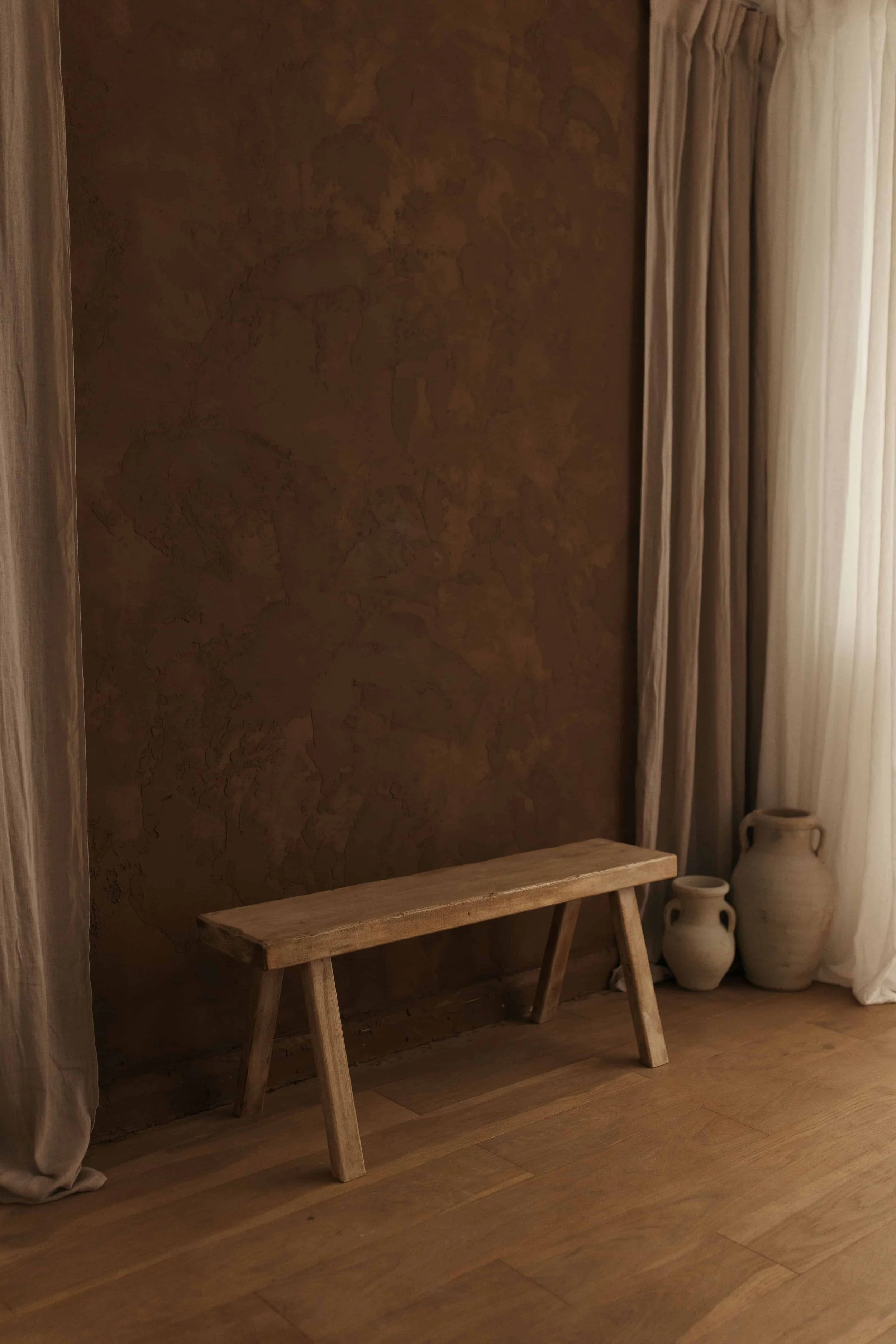 A wooden bench in front of a textured brown wall with beige curtains on the right side and several white pottery vases on the floor near the curtains.