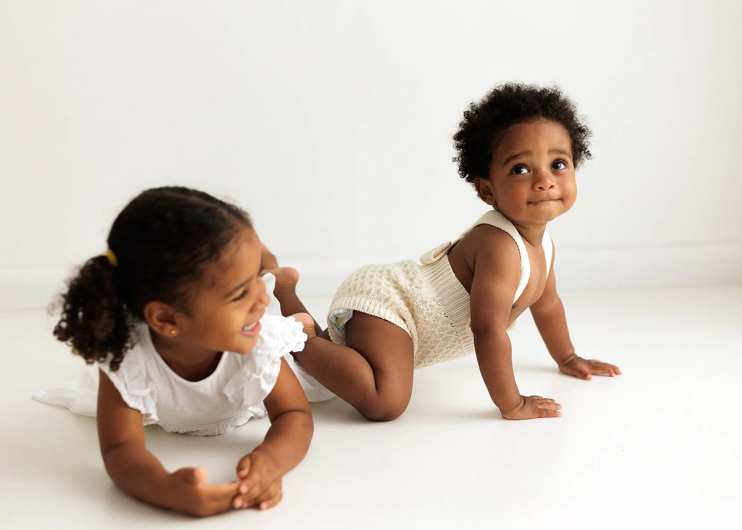 Two young children with curly hair and dark skin playing on the floor against a plain white background. One child is smiling and lying on her stomach, and the other is on her hands and knees looking upward.
