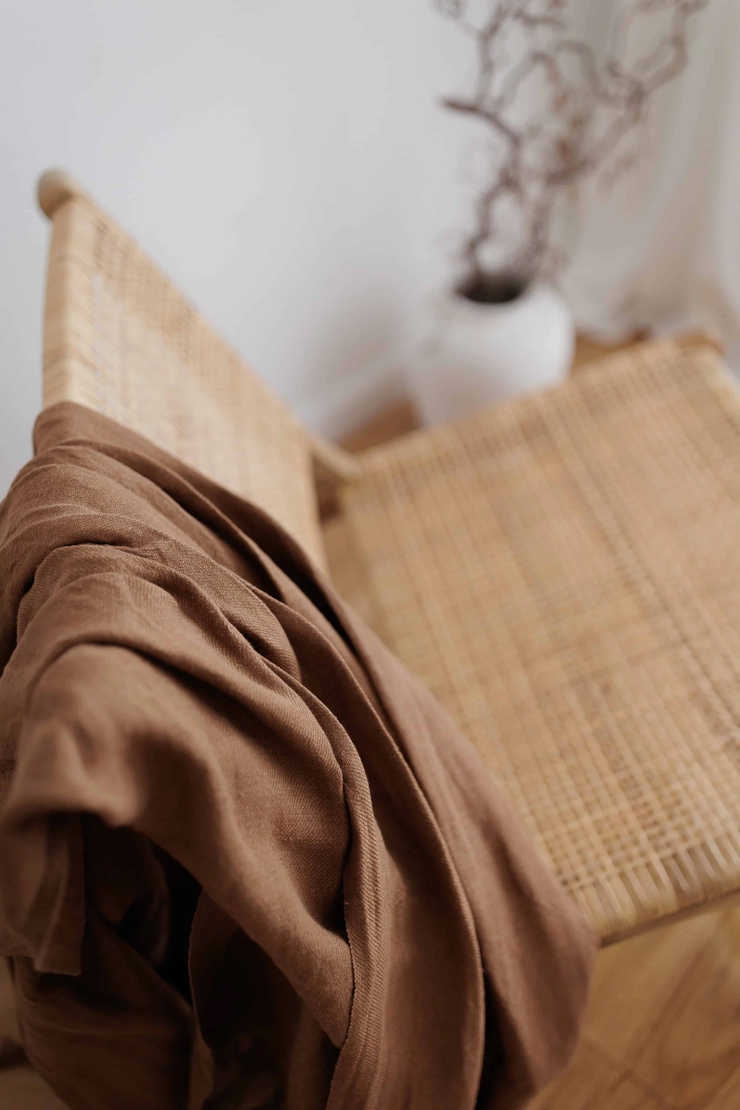 Close-up of a brown shirt draped over a wooden chair with a woven seat, with a blurry white vase containing dried branches in the background.