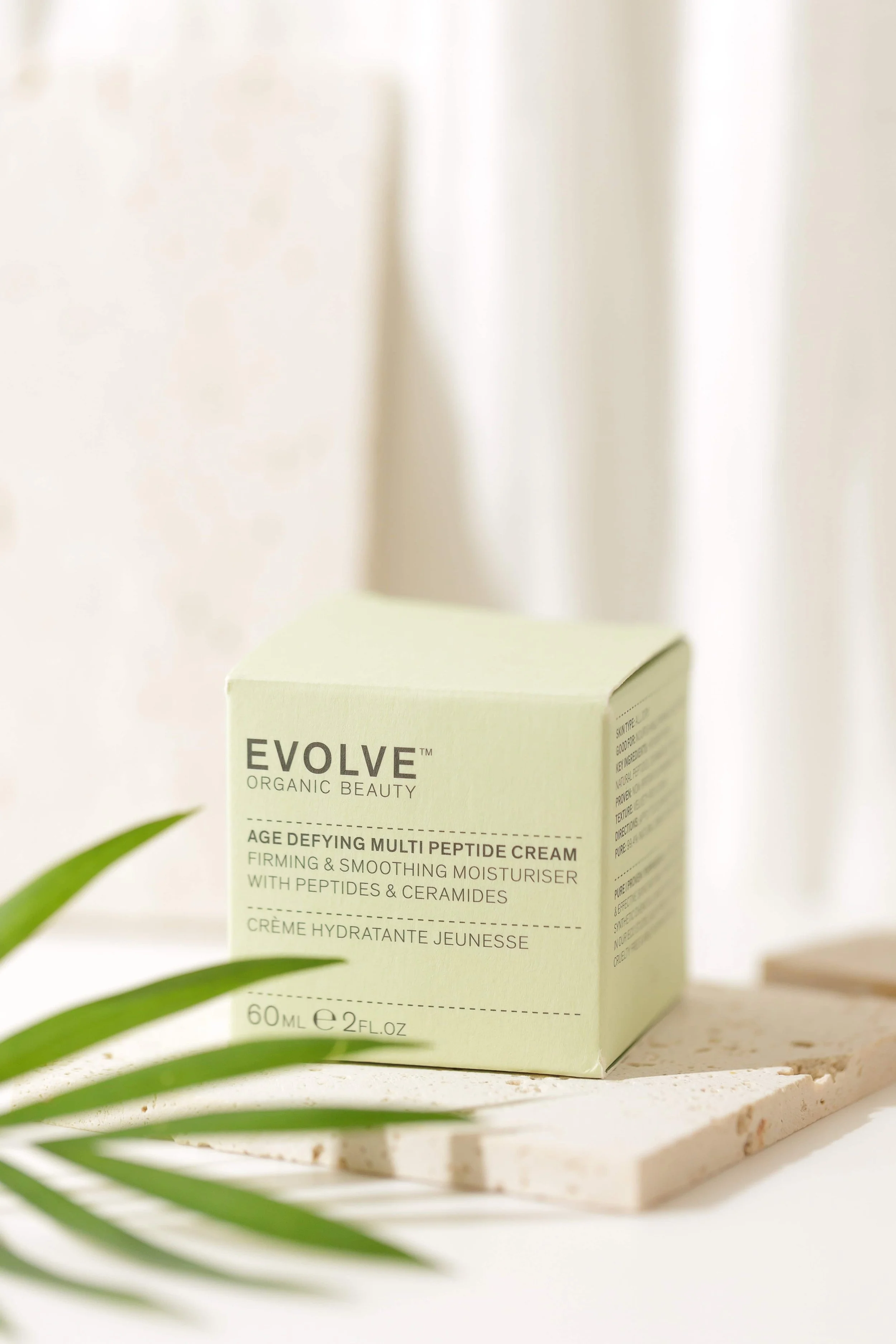 A light green box of EVOLVE organic beauty Age Defying Multi Peptide Cream on a white marble slab, with a palm leaf in the foreground and a white curtain in the background.