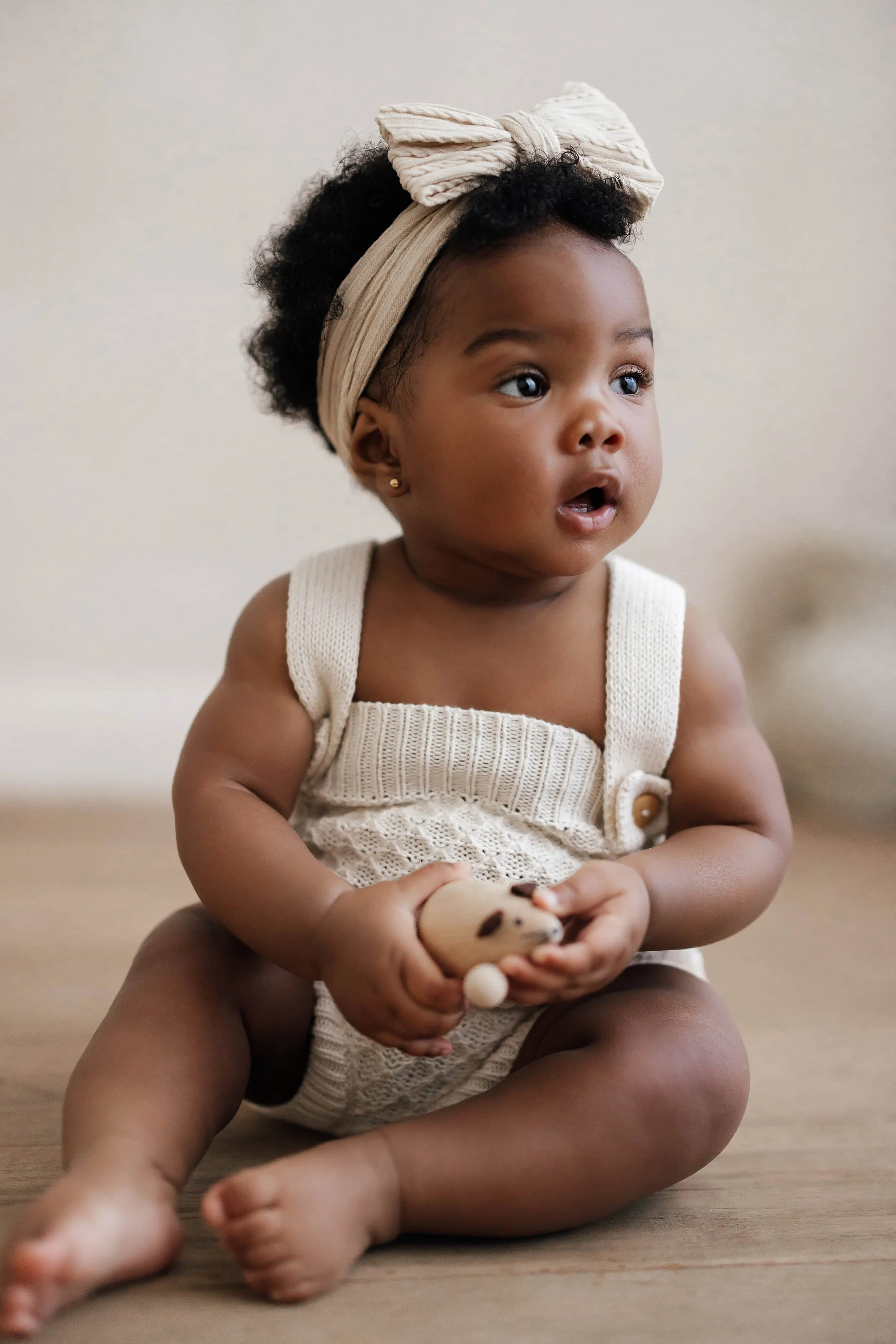 8 month old baby girl with black skin, wearing a knitted beige romper and playing with a wooden toy