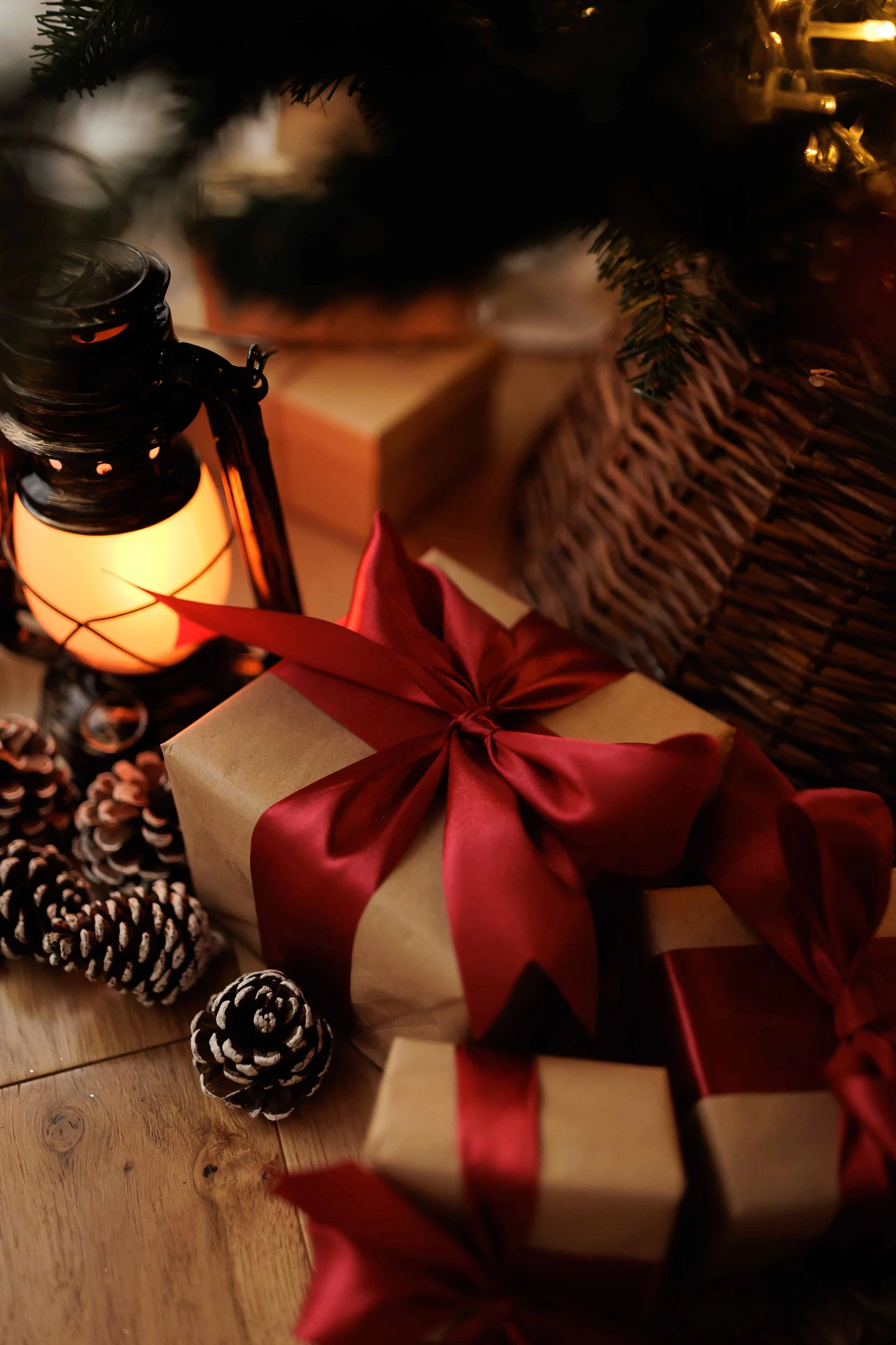 Wrapped Christmas gifts with red ribbons, pinecones, and a lantern under a decorated Christmas tree with a wicker basket nearby.