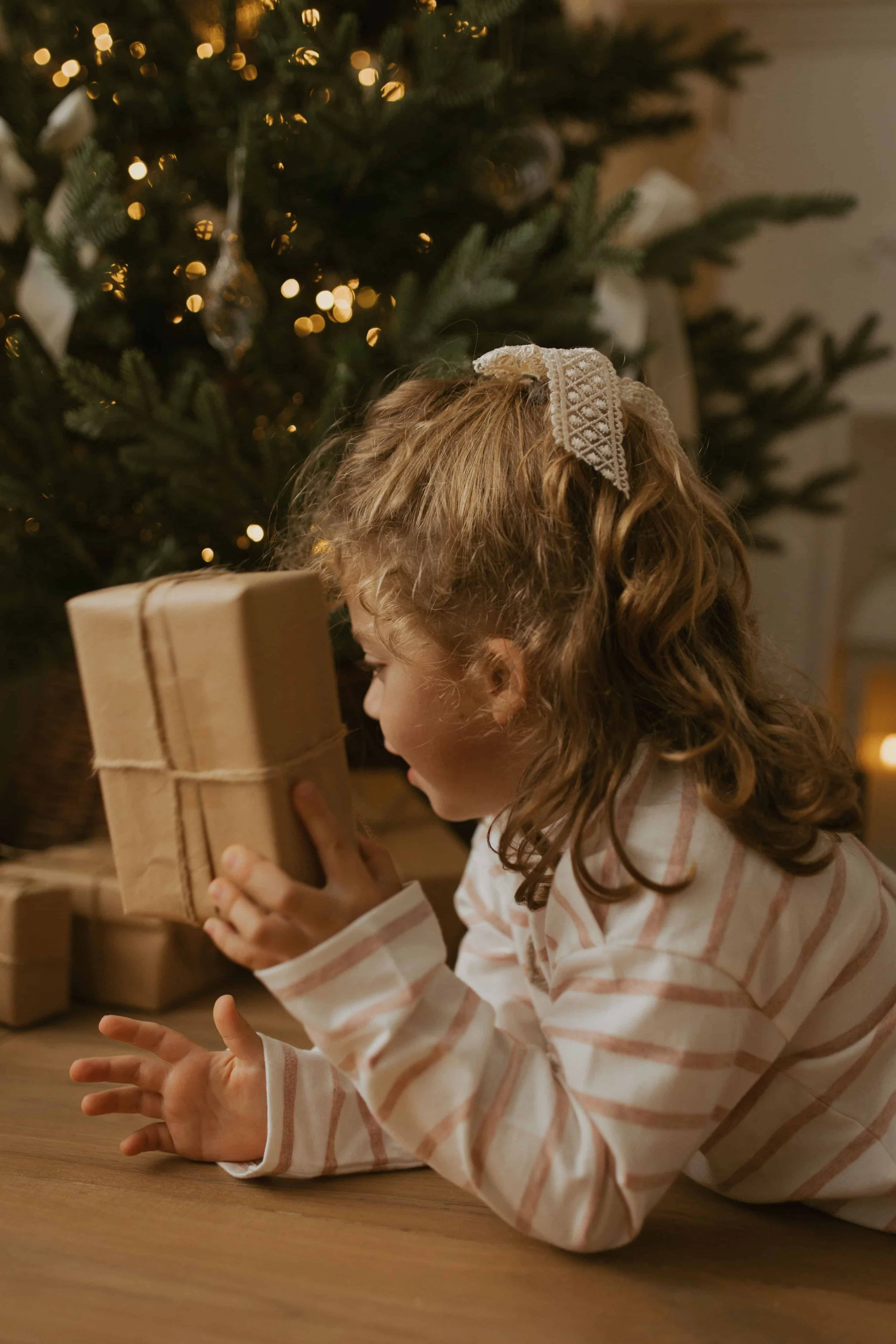 A young girl with curly red hair, wearing a striped shirt, sitting at a table and holding a wrapped gift in her hands in front of a decorated Christmas tree with lights.