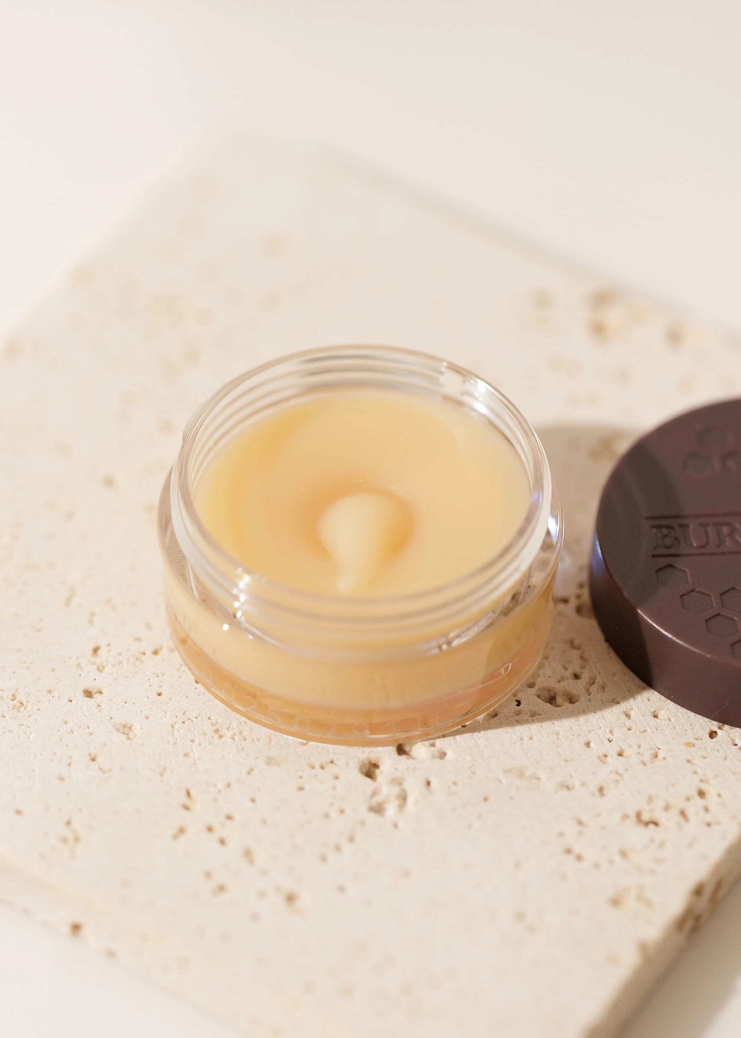 A small round container of pale yellow cream burt bee lip mask, placed on a textured beige surface next to a round dark brown lid.